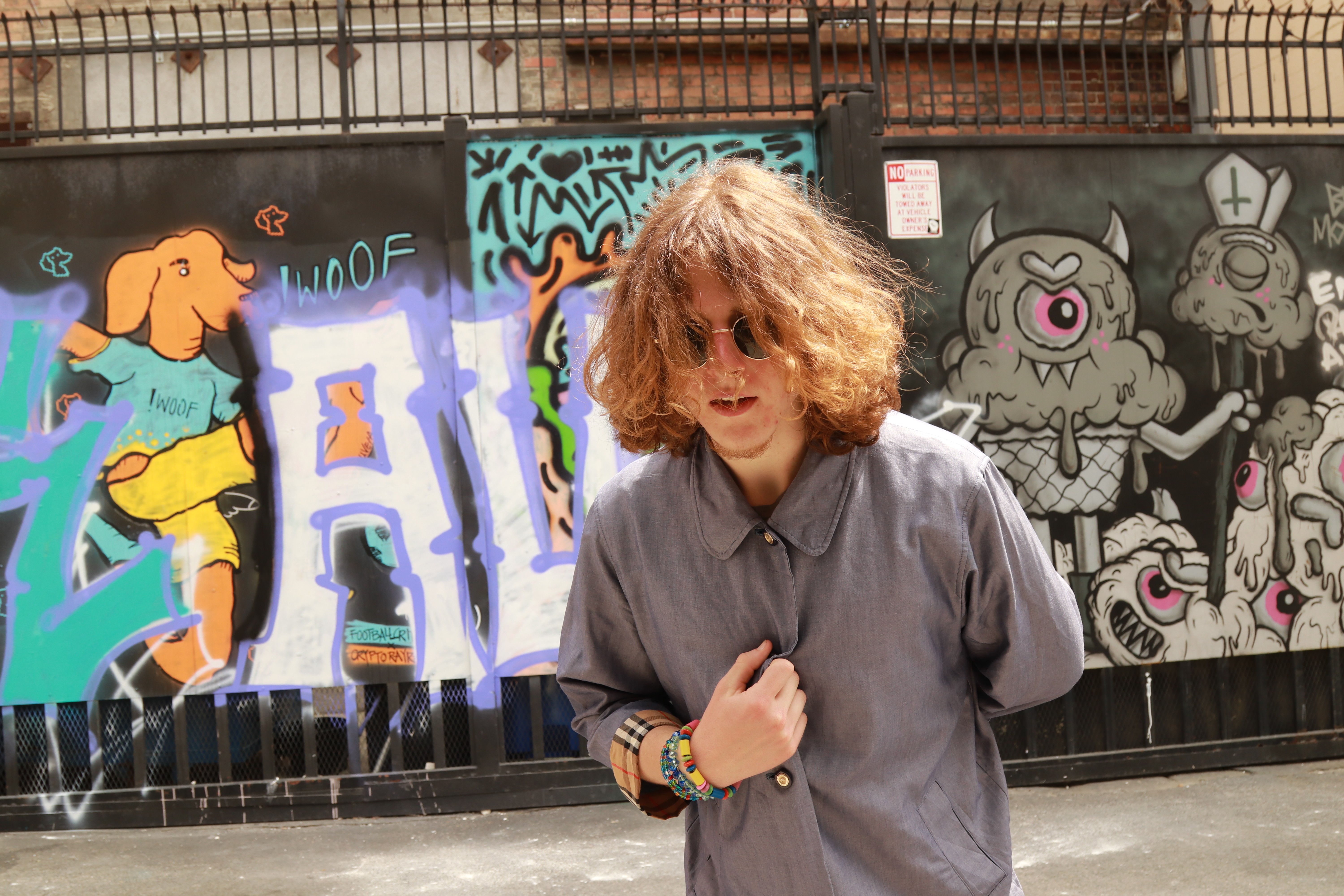 Flexer, a young music producer with curly hair and sunglasses, stands in front of a vibrant graffiti wall. Wearing a gray jacket and colorful bracelets, he strikes a confident pose. The graffiti features a cartoon dog and abstract designs, reflecting an urban and creative atmosphere.