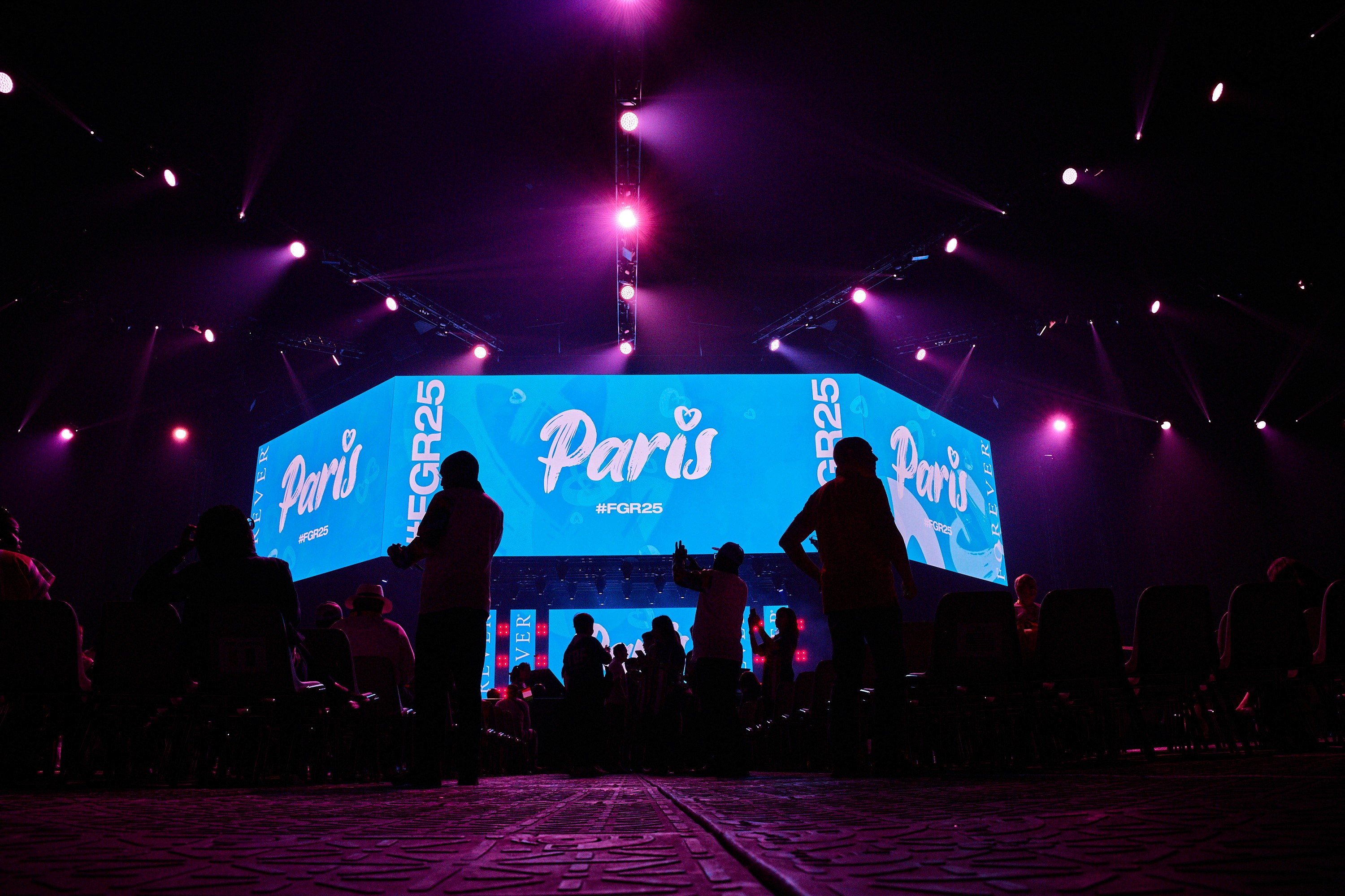 Audience silhouettes in front of a large stage with illuminated screens and lighting at a live event.