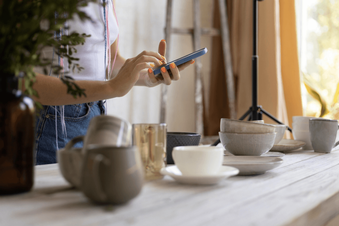 Close-up of a person using a smartphone while standing at a table with ceramic bowls and cups in a softly lit workspace.