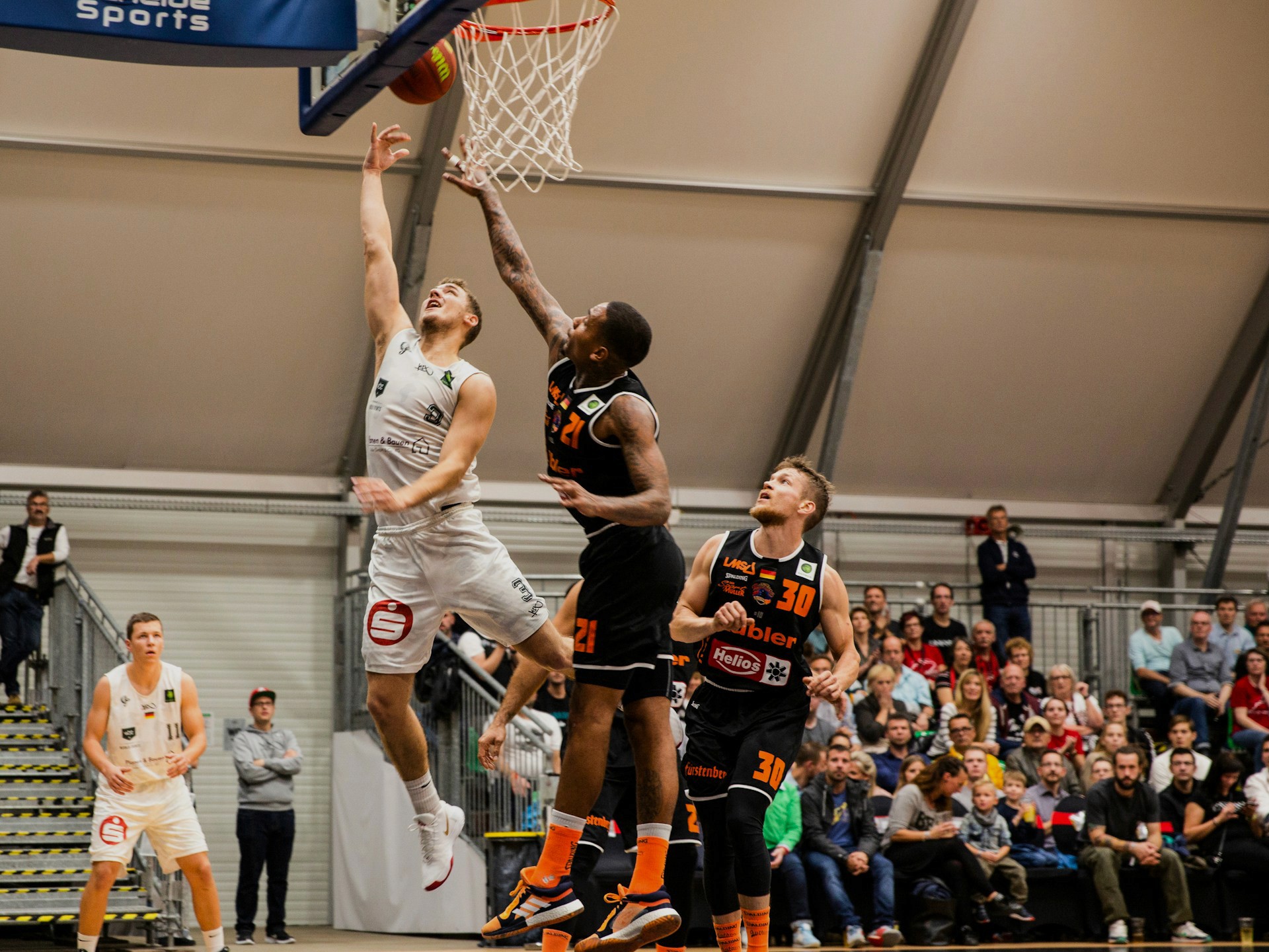 A basketball player in a white uniform jumps for a layup while a defender in a black uniform attempts to block the shot.