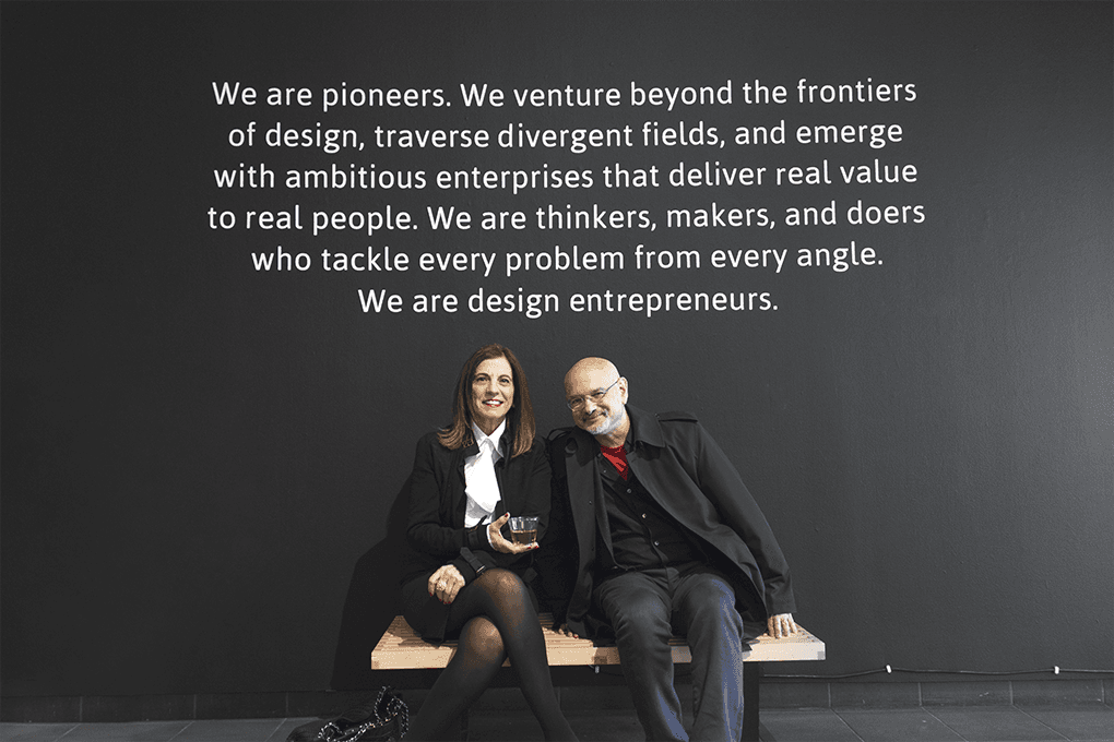 A photo of two teachers sitting on a bench. Also behind them on a wall there is a text: We are pioneers. We venture beyond the frontiers of design, travers divergent fields, and emerge with ambitious enterprises that deliver real value to real people. We are thinkers, makers, and doers who tackle every problem from every angle. We are design entrepreneurs.
