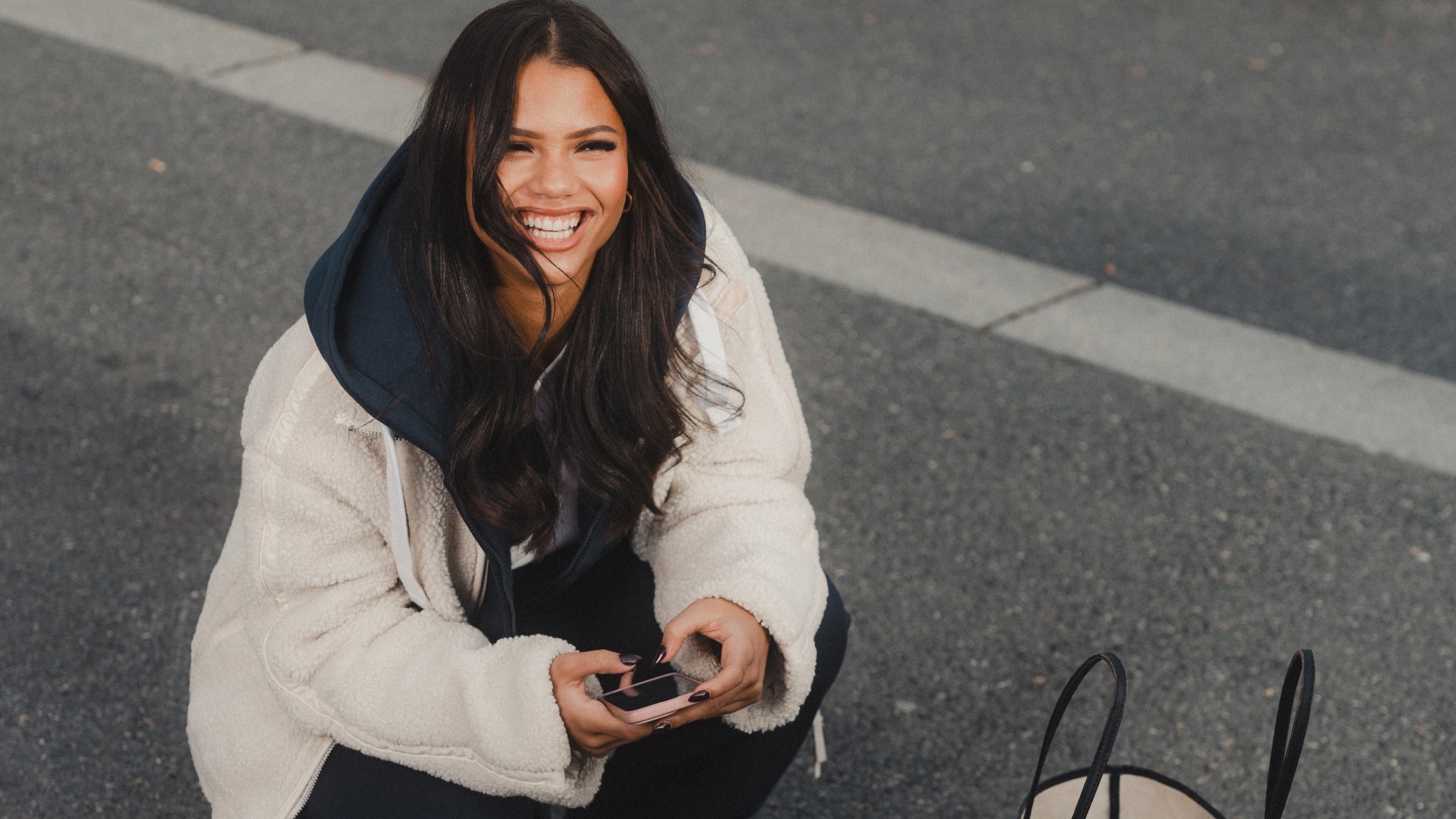 Young woman dressed in dark blue slacks and a fluffy white jacket sitting on the asphalt in an urban area