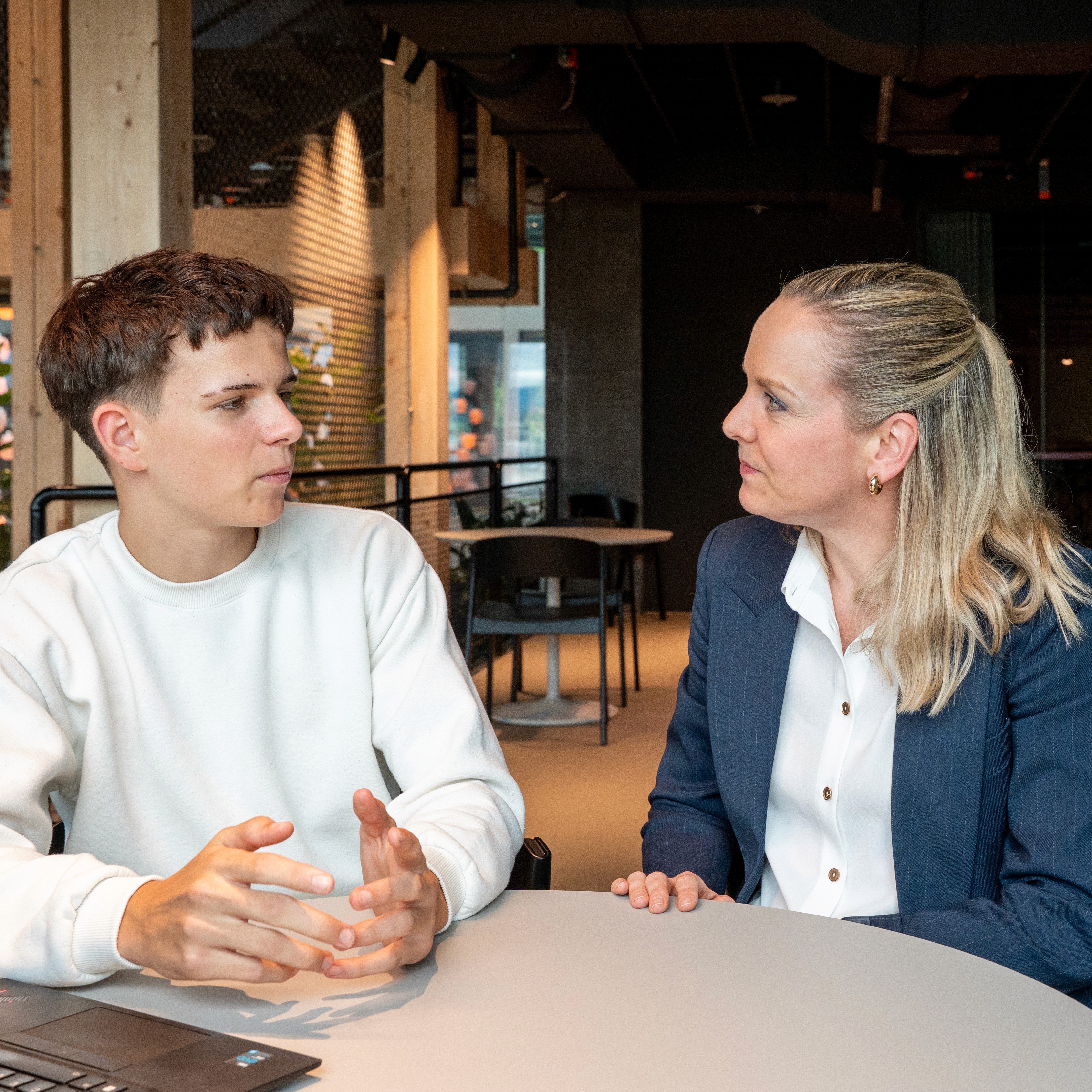 A young person with curly hair smiles at the camera while sitting in front of a laptop against a dark background.