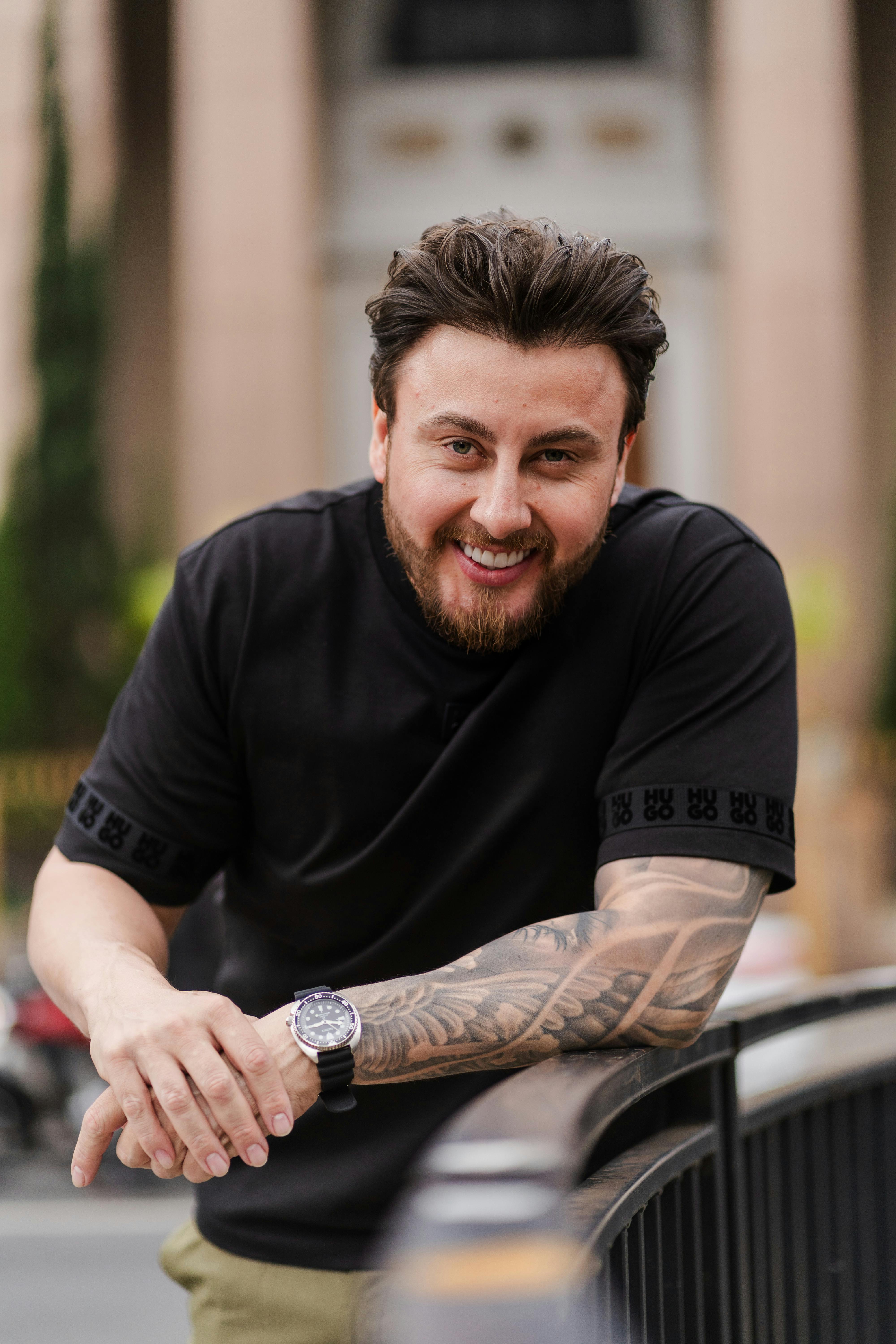 A smiling man with tattoos leans casually on a car, posing outdoors in a relaxed setting.