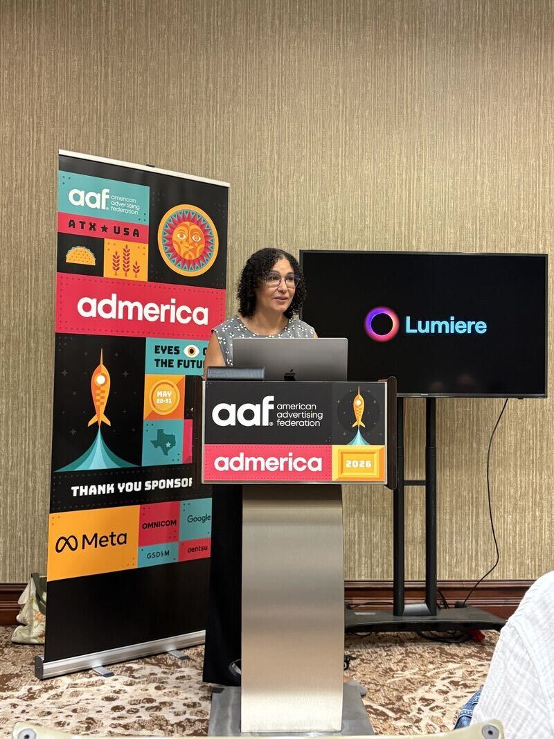 Amber Williams, COO, speaks at a podium with the American Advertising Federation 'AdAmerica 2026' branding, standing beside a colorful AAF AdAmerica banner and a screen displaying the Lumiere logo