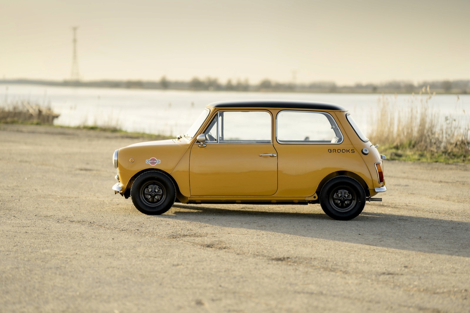 A vintage orange car parked on a road with a scenic background of calm water and gentle hills.