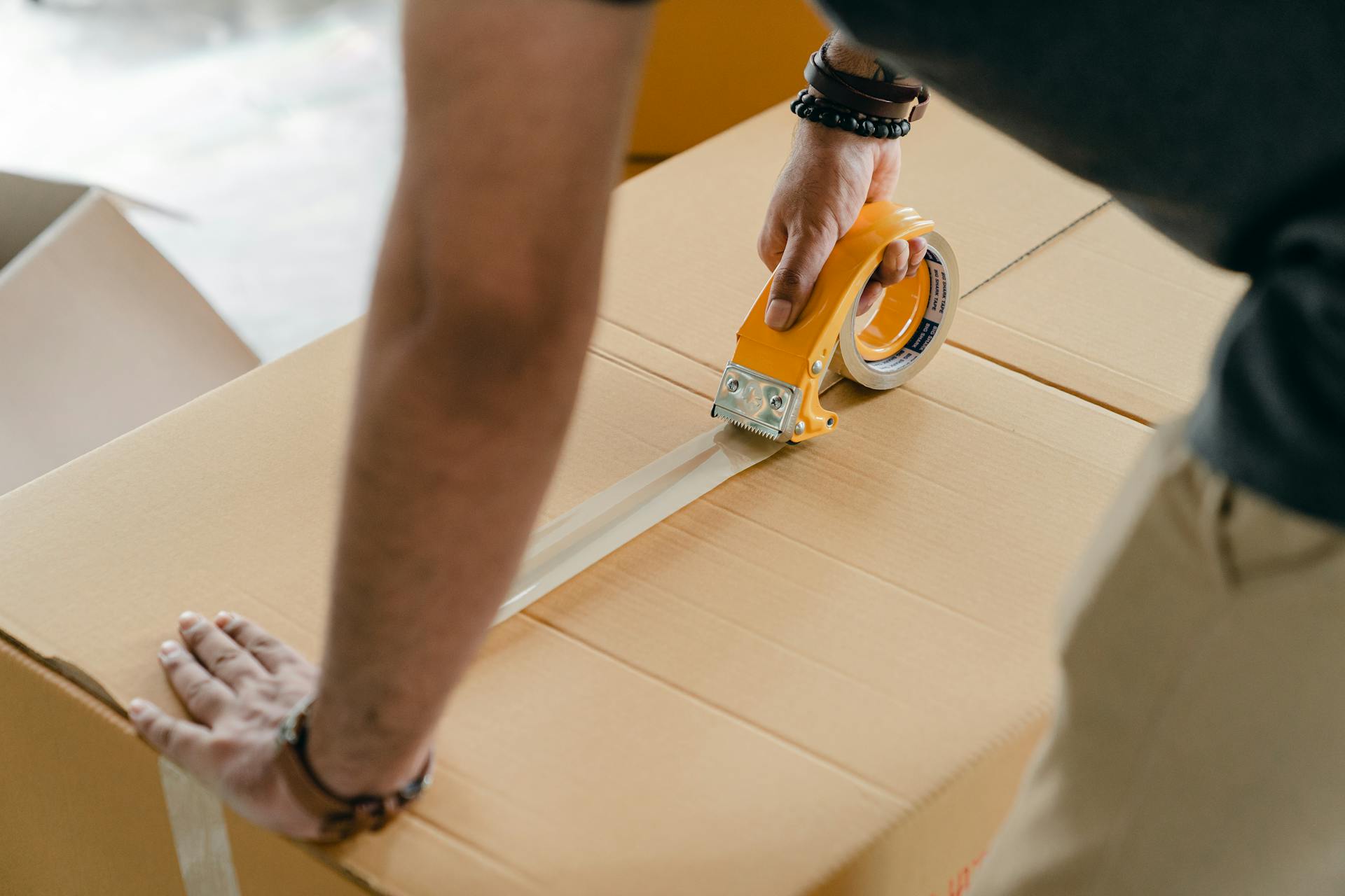 A close-up shot of a person using a handheld yellow tape dispenser to seal the top of a brown cardboard box with clear packing tape. The person is wearing bracelets and has their hand on the box to stabilize it while taping.