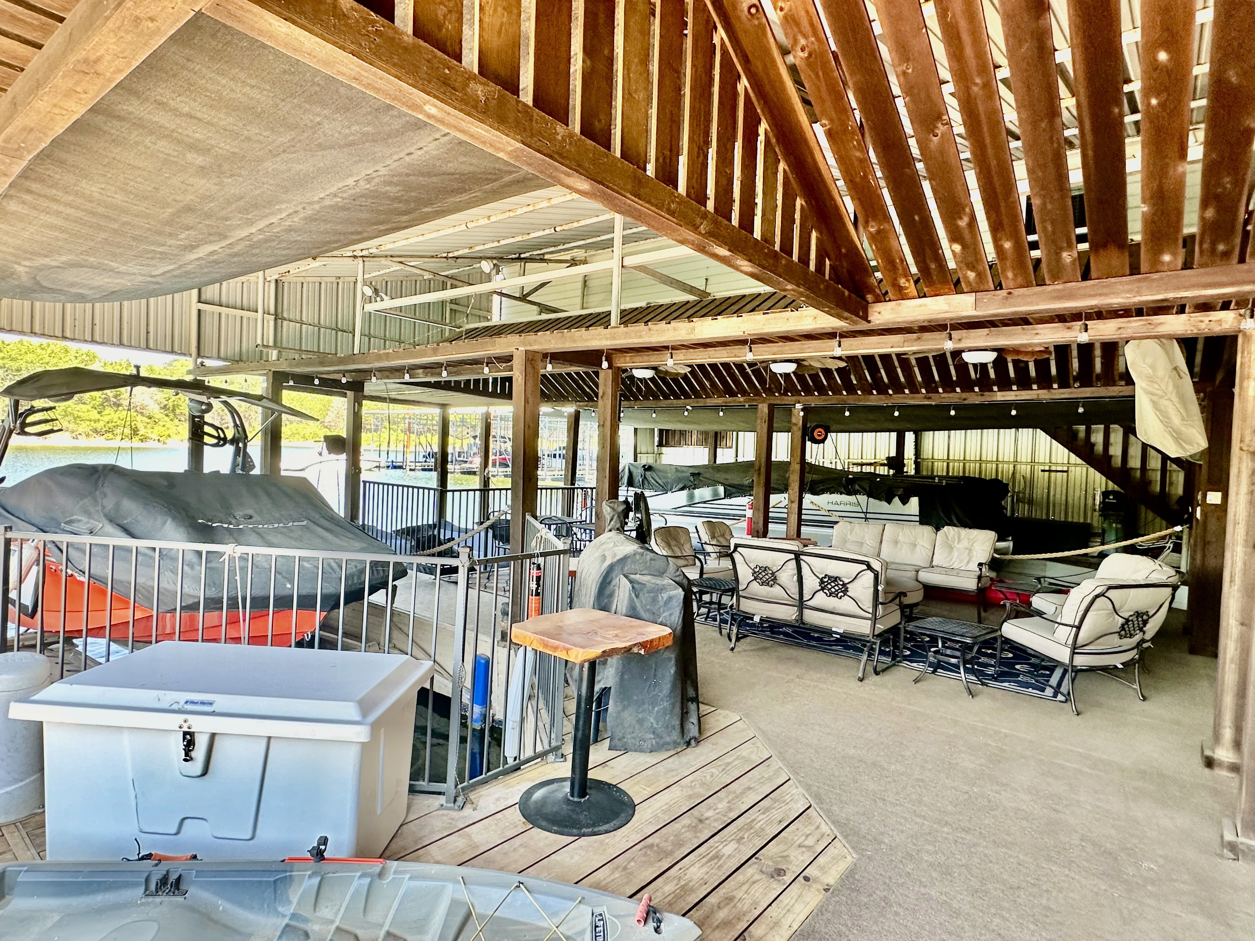 The covered boat dock features a seating area with cushioned patio furniture, overlooking boats stored under a wooden-beamed roof.