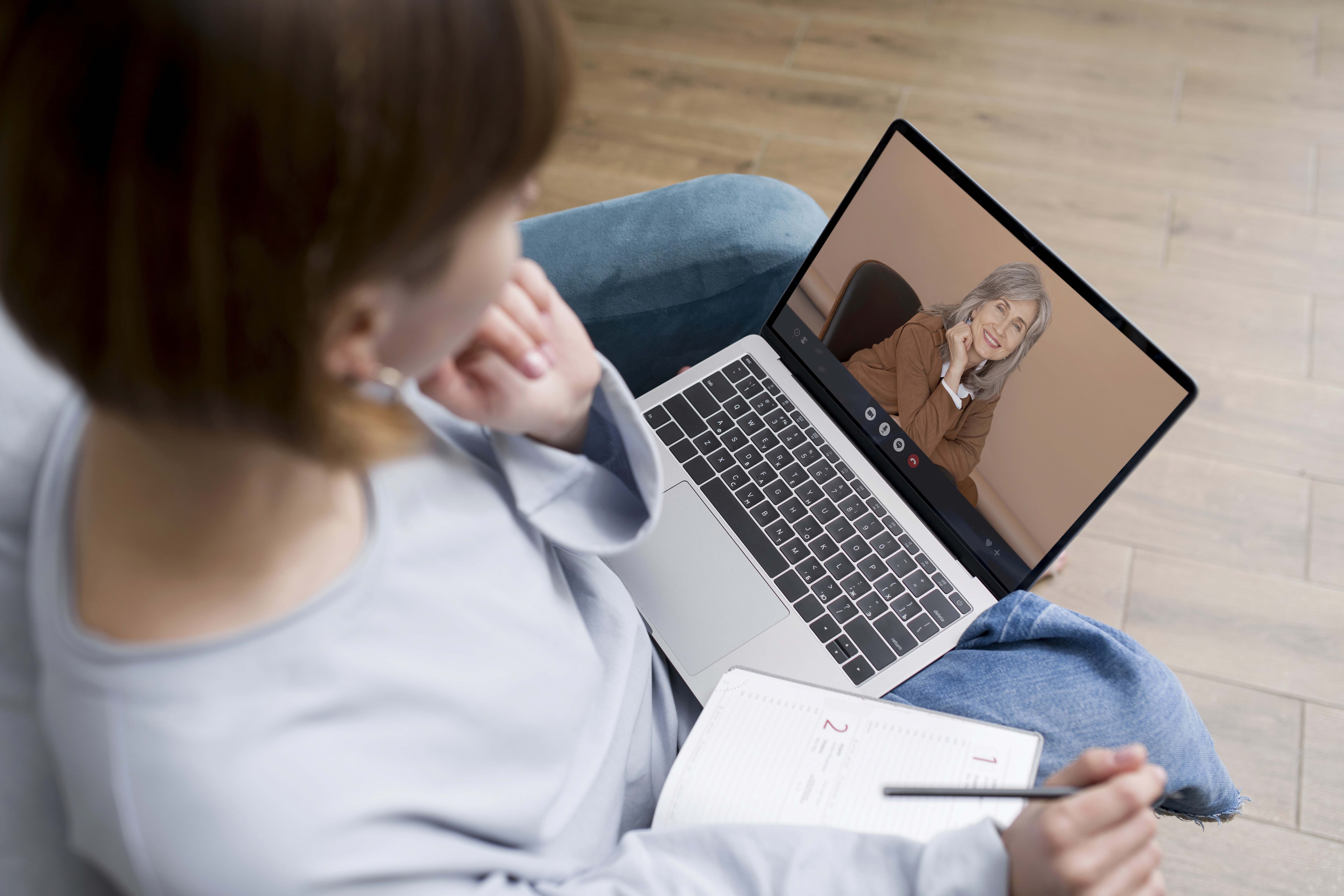 Eine Frau sitzt auf einem Stuhl und hat ihren Laptop auf dem Schoß. Sie hat einen Video-Call mit einer Beraterin und macht sich nebenbei Notizen..