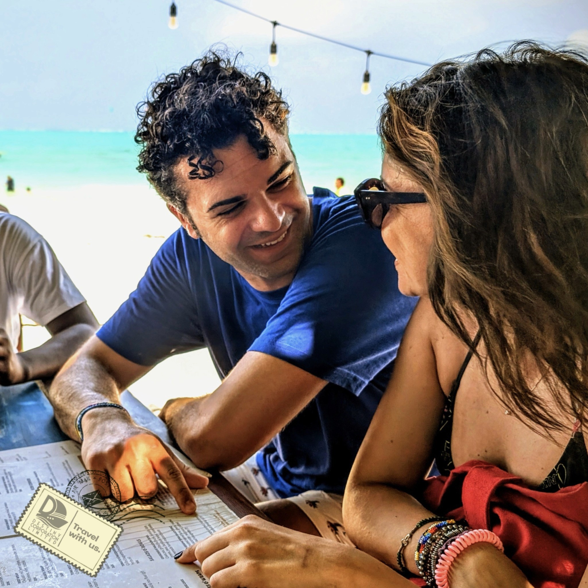 Group of young travelers sitting together in AlUla’s old town, sharing drinks and stories under palm trees on a sunny day.