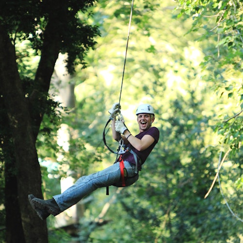 Persona che fa zipline attraverso una foresta lussureggiante e verde, sorridente e indossando equipaggiamento di sicurezza, inclusi casco e imbracatura.