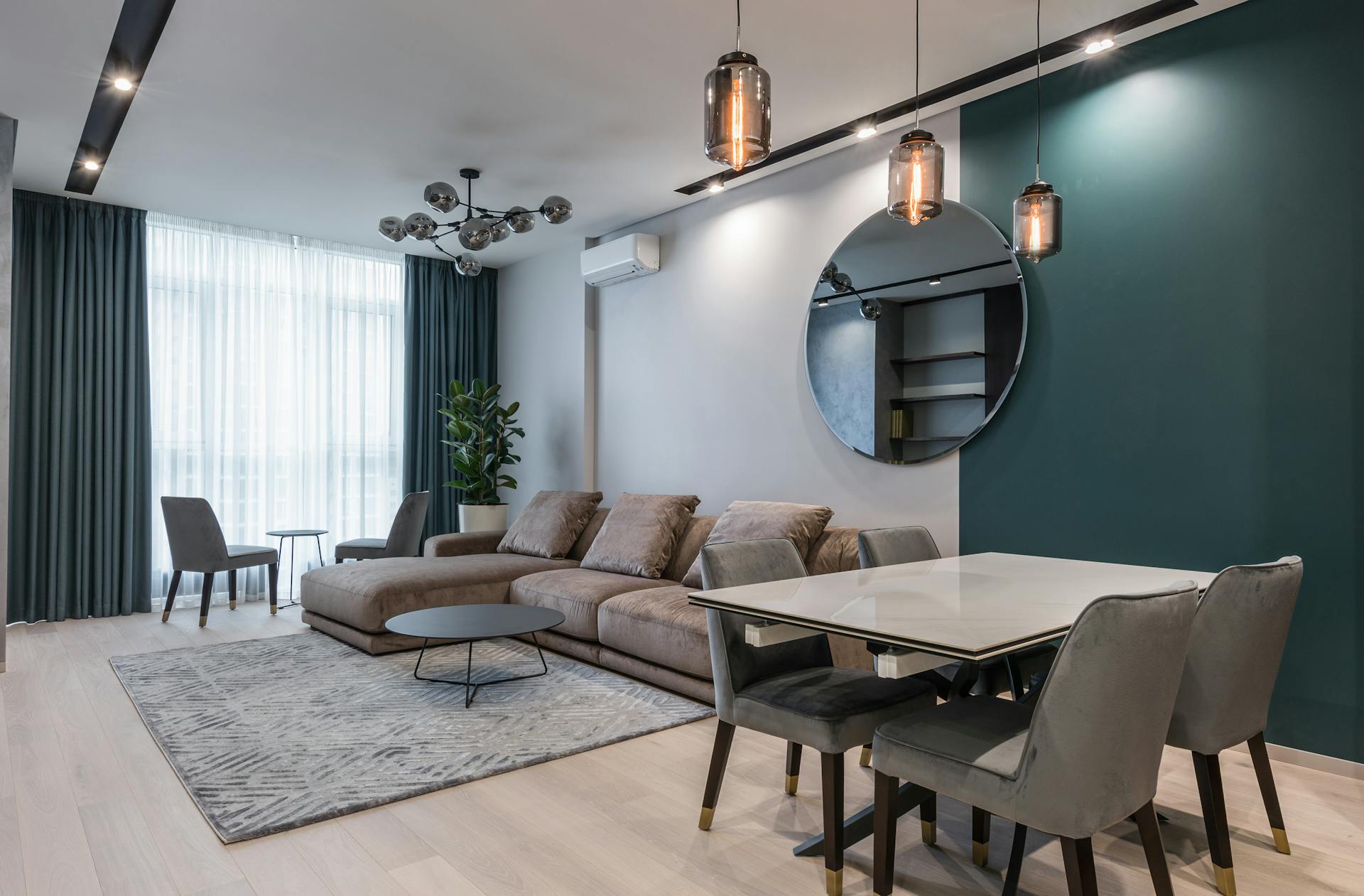 Modern open-plan living and dining room with a large taupe sectional sofa, a white marble dining table, gray velvet chairs, a round mirror, and a dark teal accent wall.