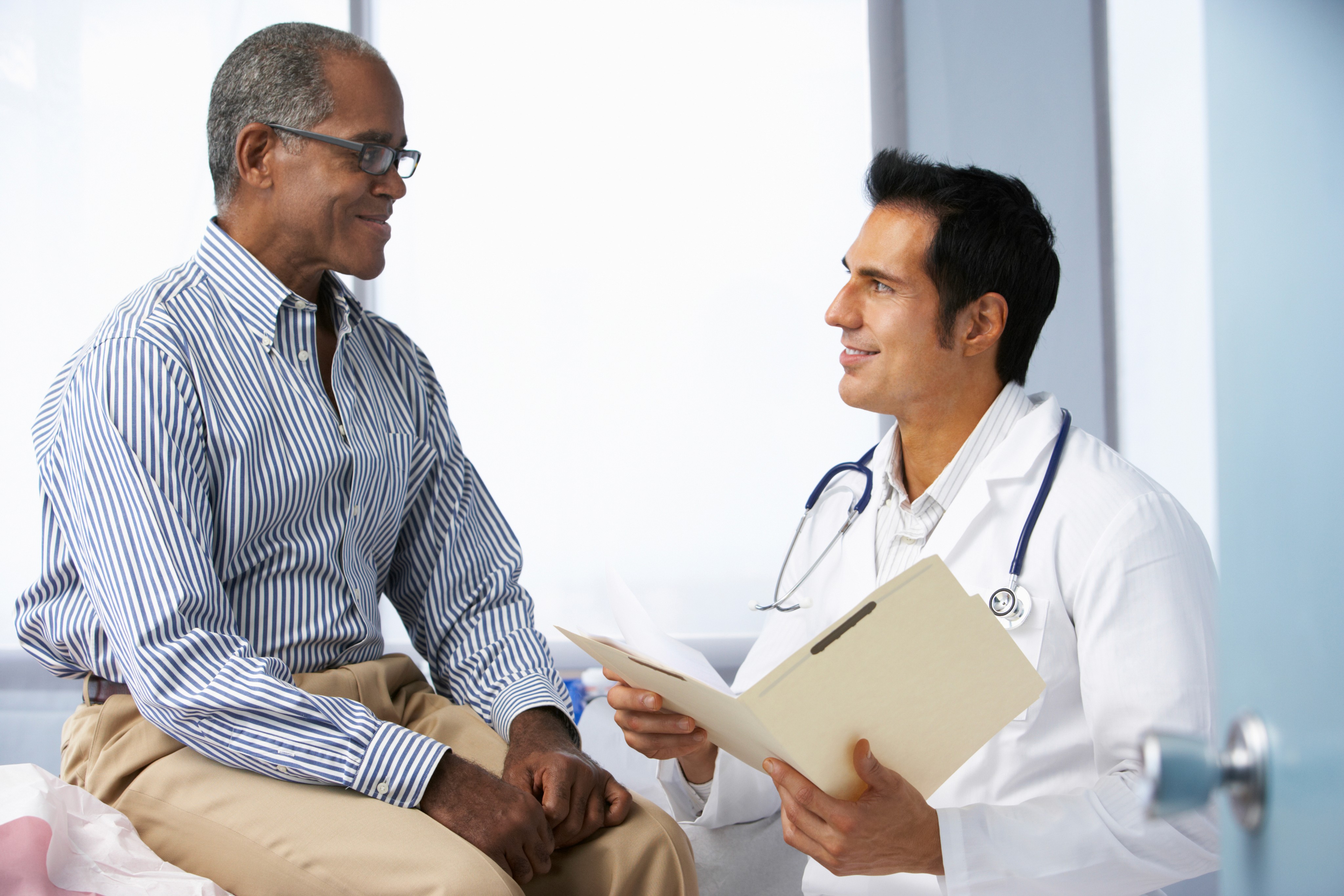 Doctor in white coat holding a folder consulting with a seated older male patient in a medical office.