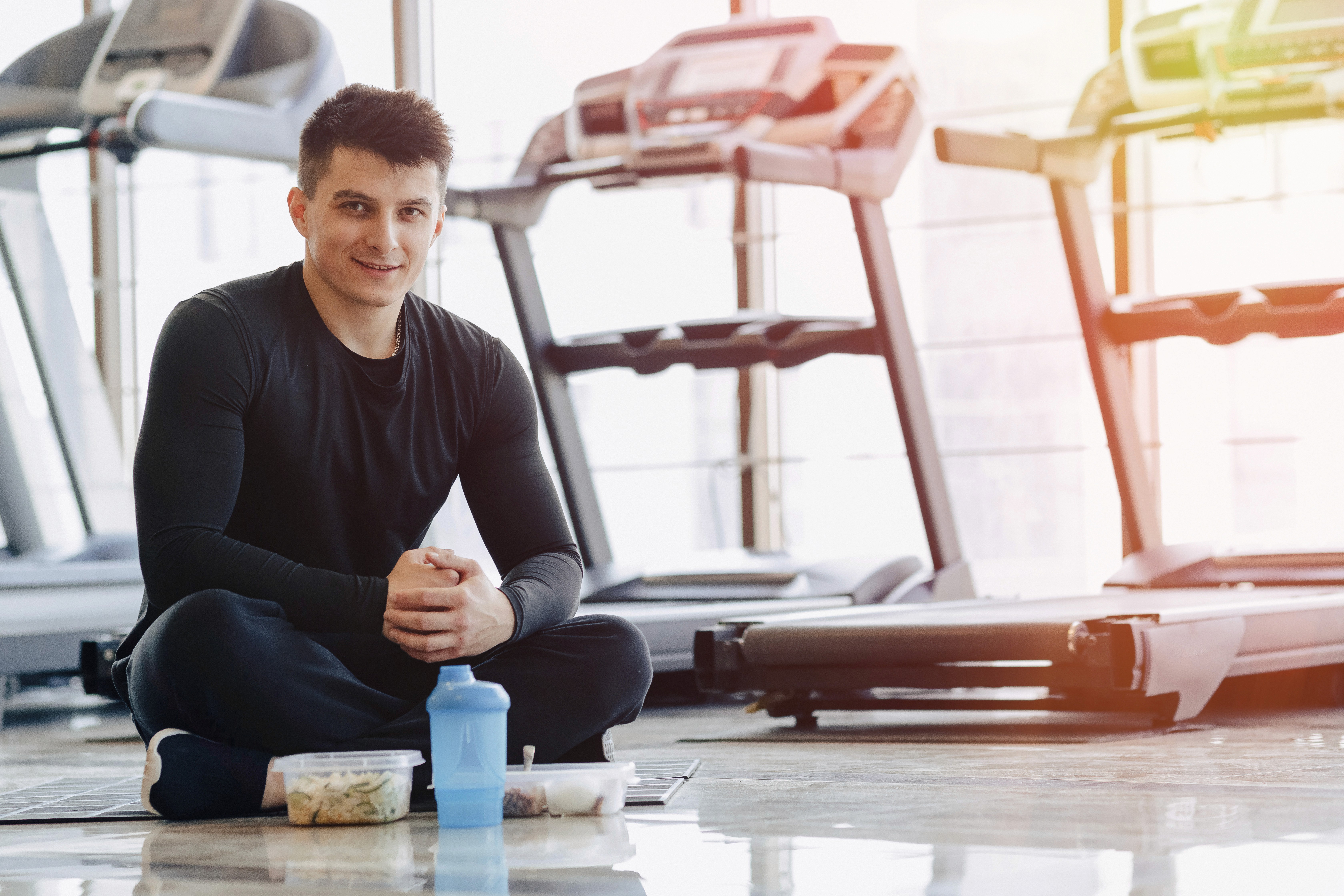 Man in black workout clothes sits cross-legged on a gym floor with a shaker bottle and meal prep containers,