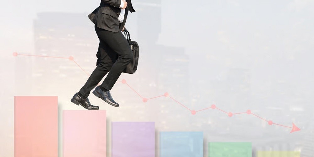 A person in a business suit carrying a briefcase, mid-stride across a series of descending bar graph columns. A red line graph with a downward arrow is overlaid in the background, symbolizing a decline or crisis, while the person moves forward against the trend