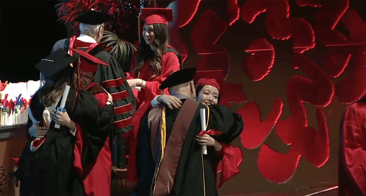 A photo of a couple of grad students and teachers, dressed in SVA color robes, that are hugging and congratulating each other.