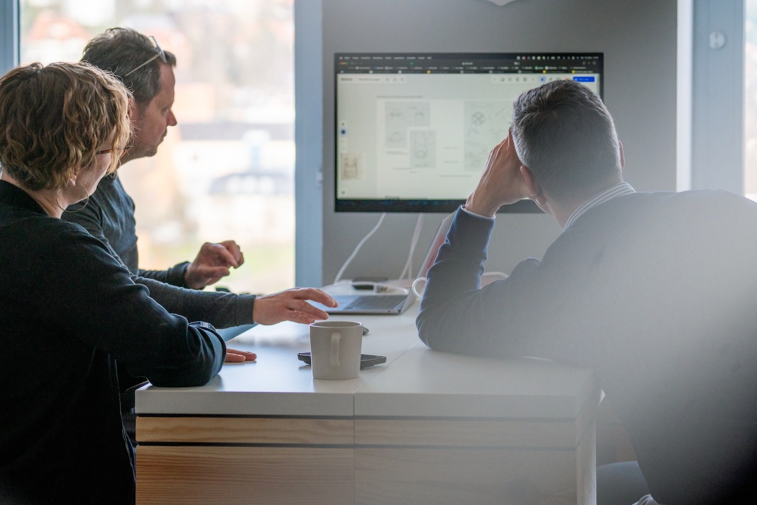 Drei Personen sitzen an einem Tisch und schauen auf einen großen Bildschirm mit geöffnetem Computerprogramm