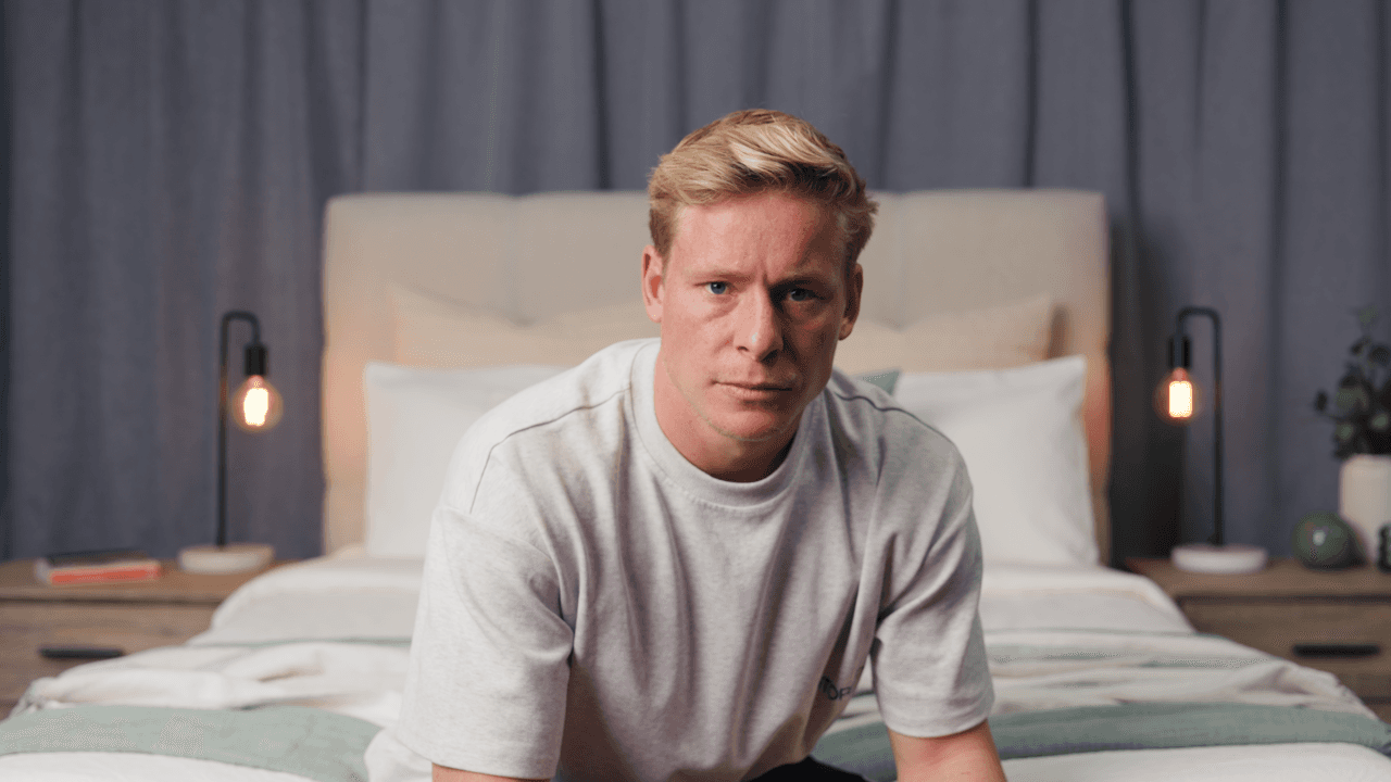 A person with blonde hair sits on a bed wearing a light-colored sweatshirt, looking at something off-camera