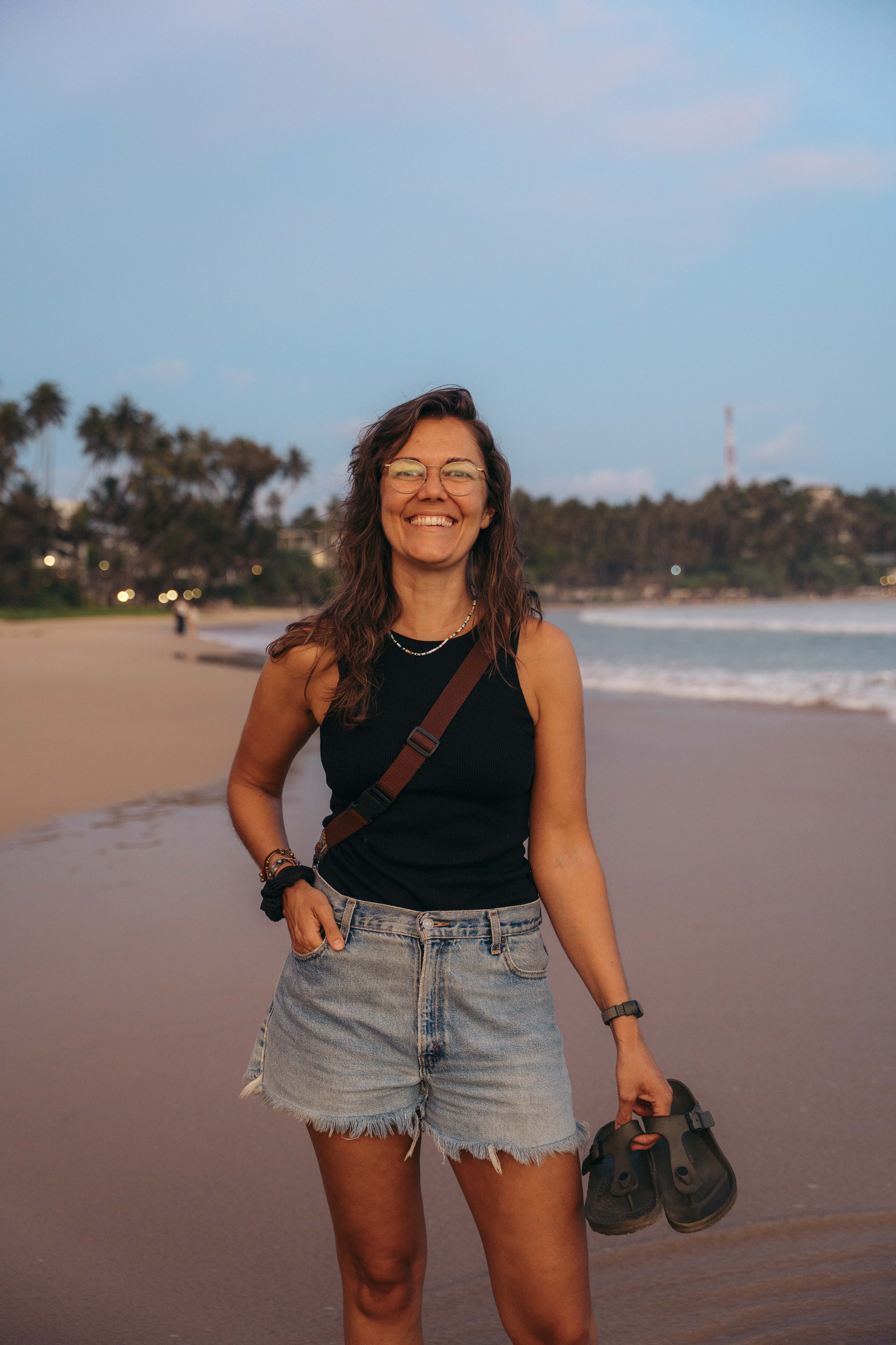 Eva smiling while walking on a sandy beach beside clear blue water.