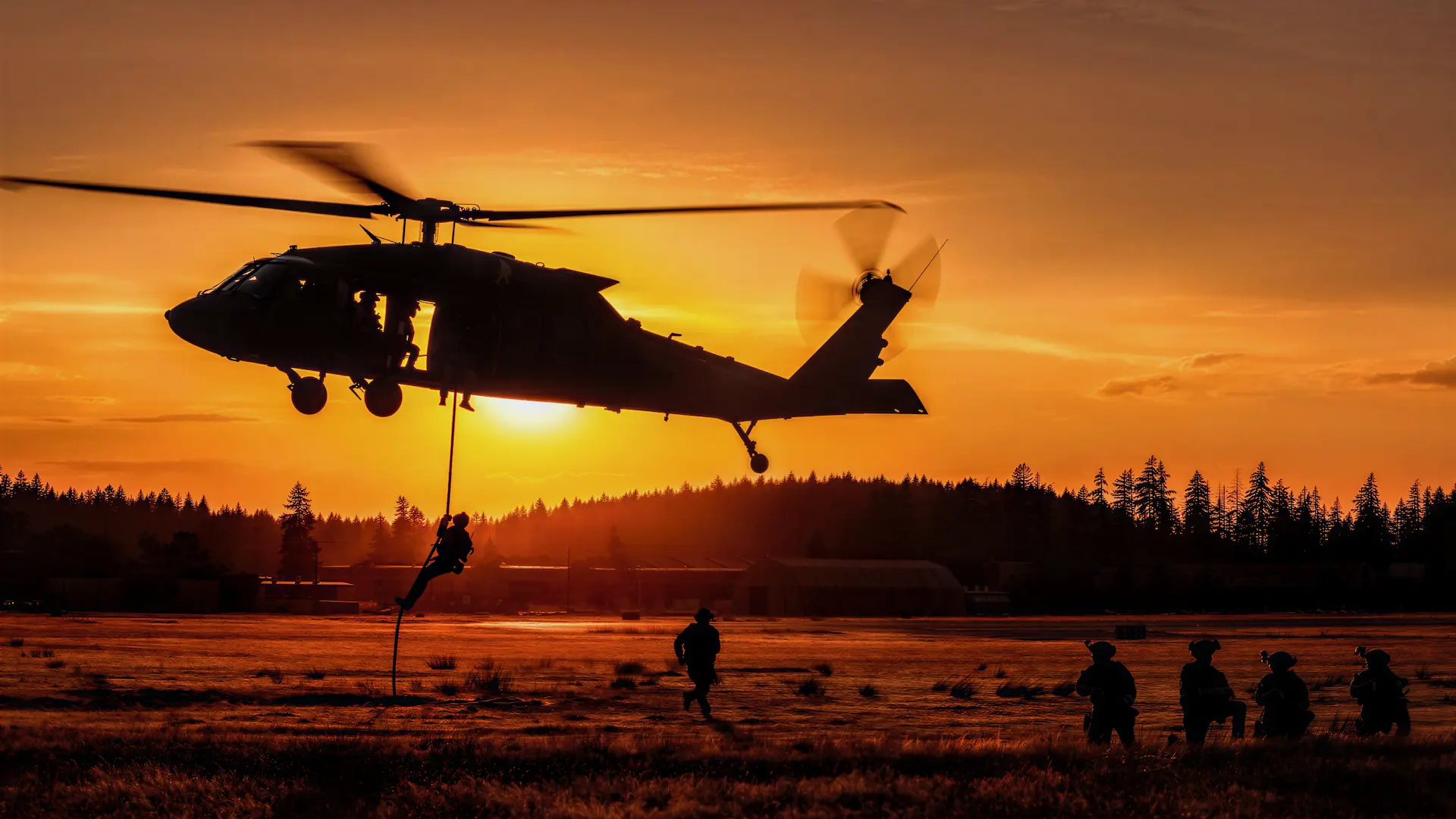 Silhouetted soldier rappels from a Black Hawk helicopter at sunset as troops move below