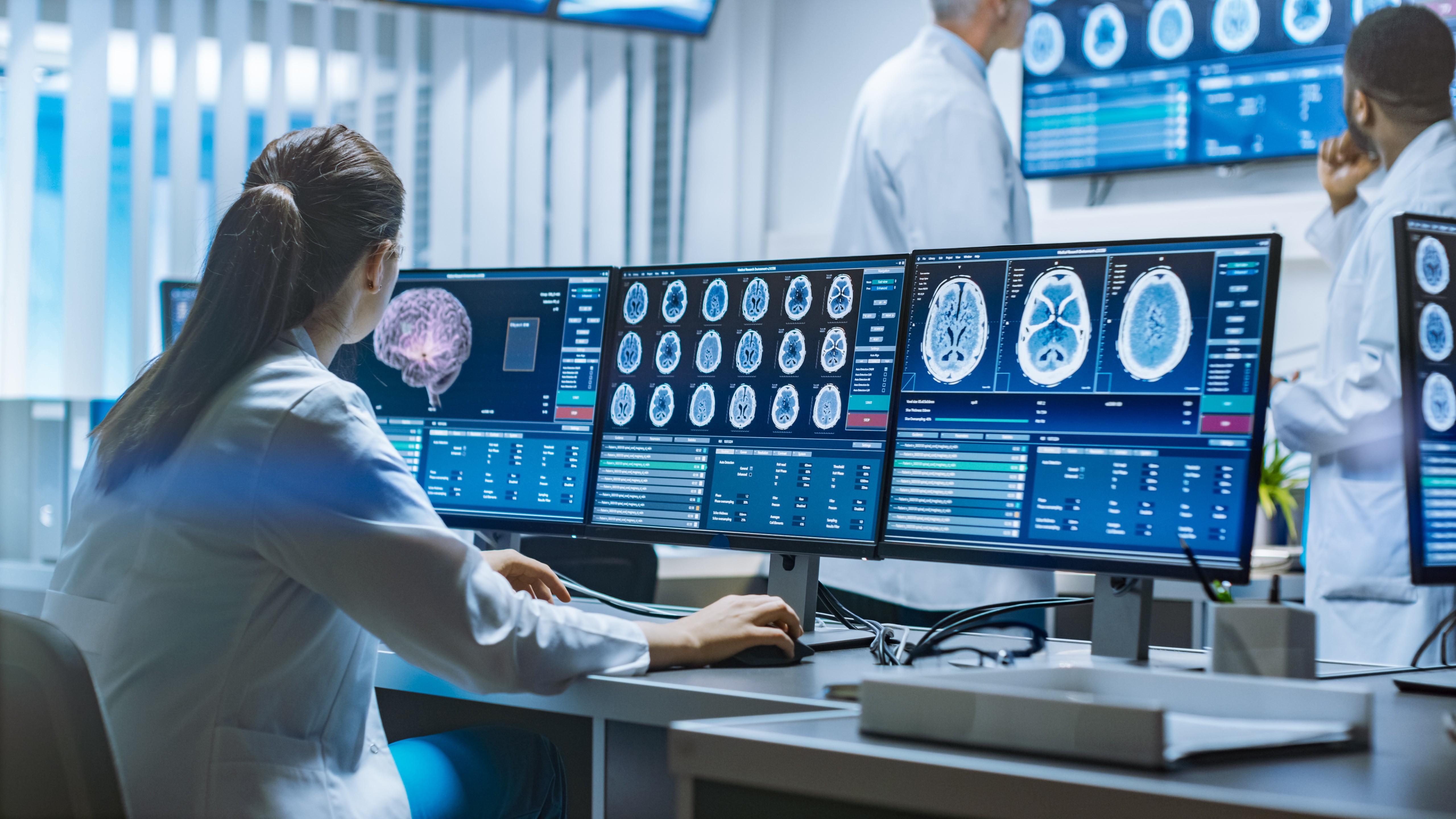 Researcher in a white coat viewing brain scan images across multiple monitors in a medical imaging lab, with colleagues nearby