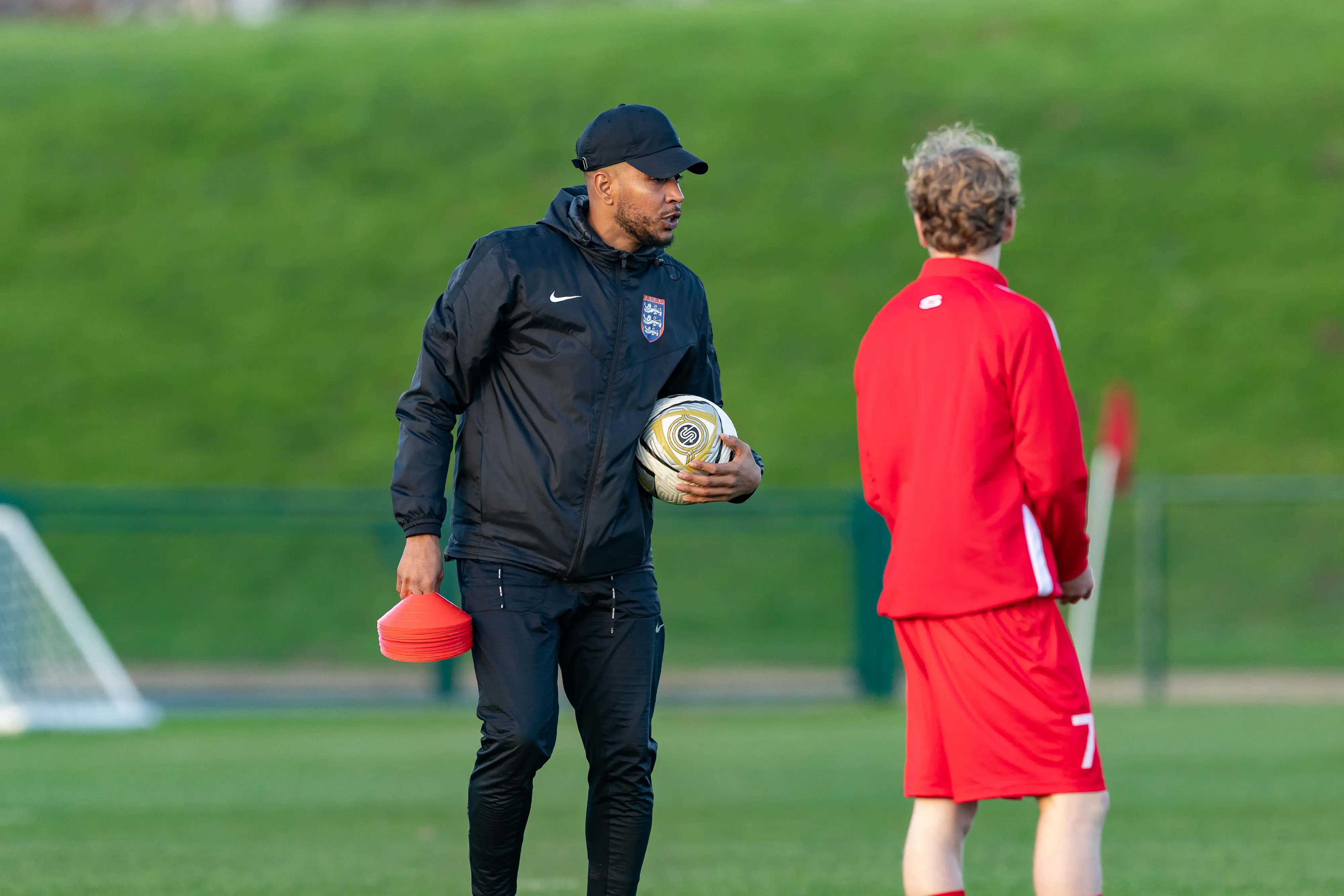 A soccer coach in black sportswear and a cap is holding a ball and red cones, speaking to a player in a red kit on a grassy field.
