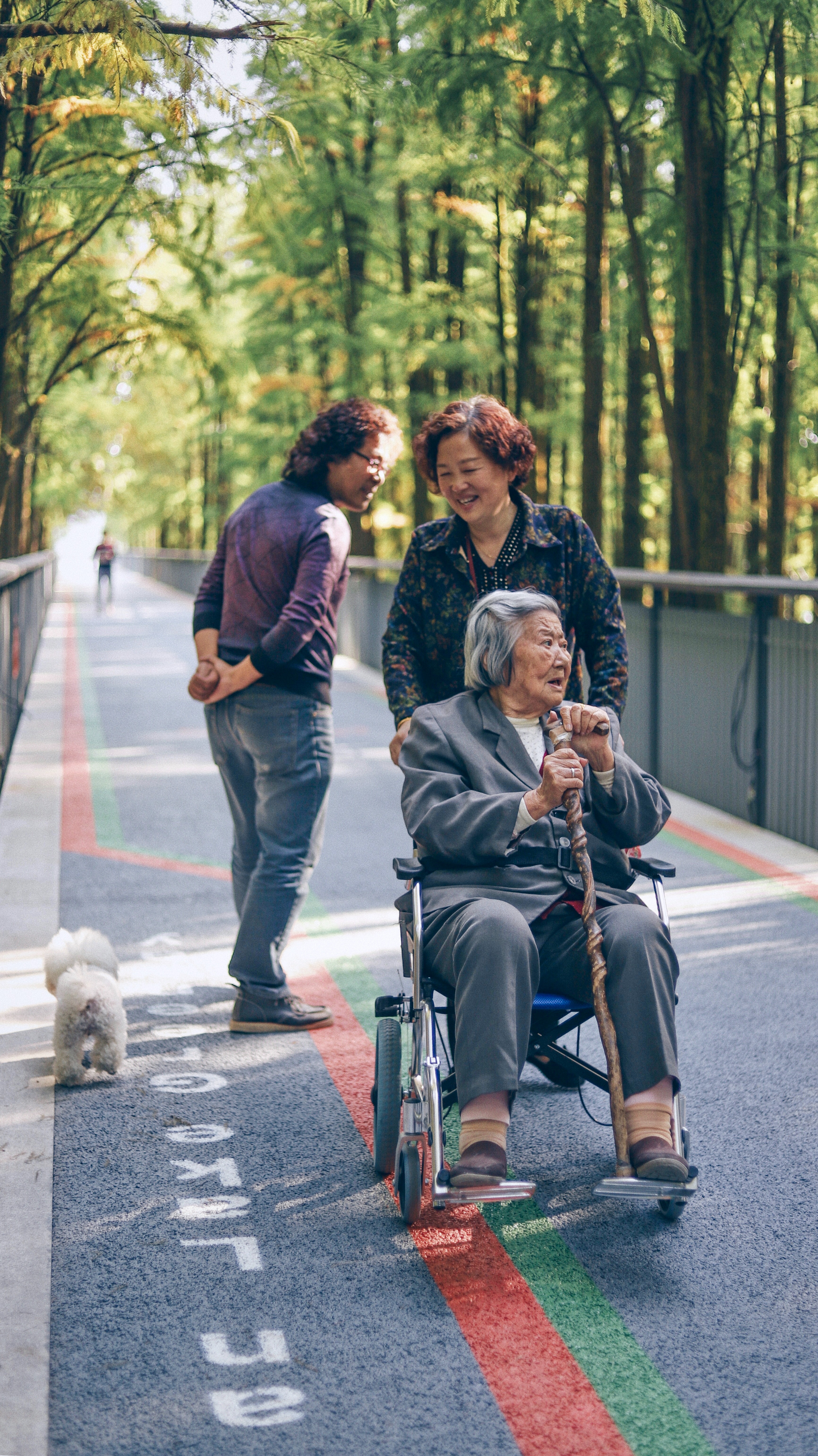 An elderly Asian woman with grey hair sits in a wheelchair outdoors, gripping a wooden cane with both hands. She looks off to the side with an expression of engagement and curiosity. Behind her, a caregiver in a floral blouse smiles down at her, suggesting a warm and attentive relationship. In the background, a blurred figure walks away, which adds to the sense of activity and life in the setting.