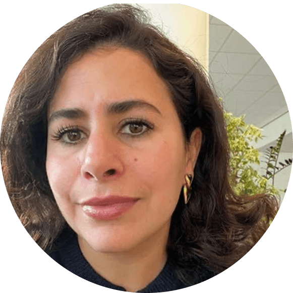 A person with long blond hair smiles warmly while standing in front of a vibrant yellow wall, wearing a black jacket and a gray turtleneck sweater.