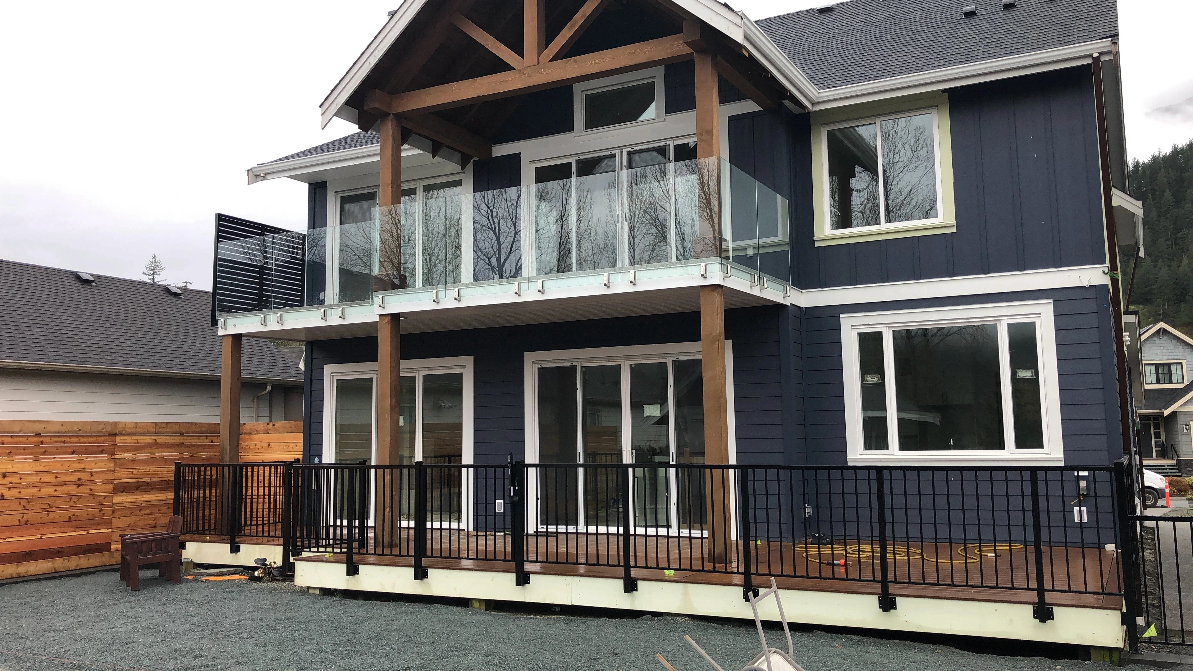 Exterior view of a modern two-story dark blue house with white window trim and prominent natural wood pillars. The upper balcony is fitted with a clear, frameless glass railing system, while the large ground-level wooden deck is fully enclosed by a black metal picket safety fence.