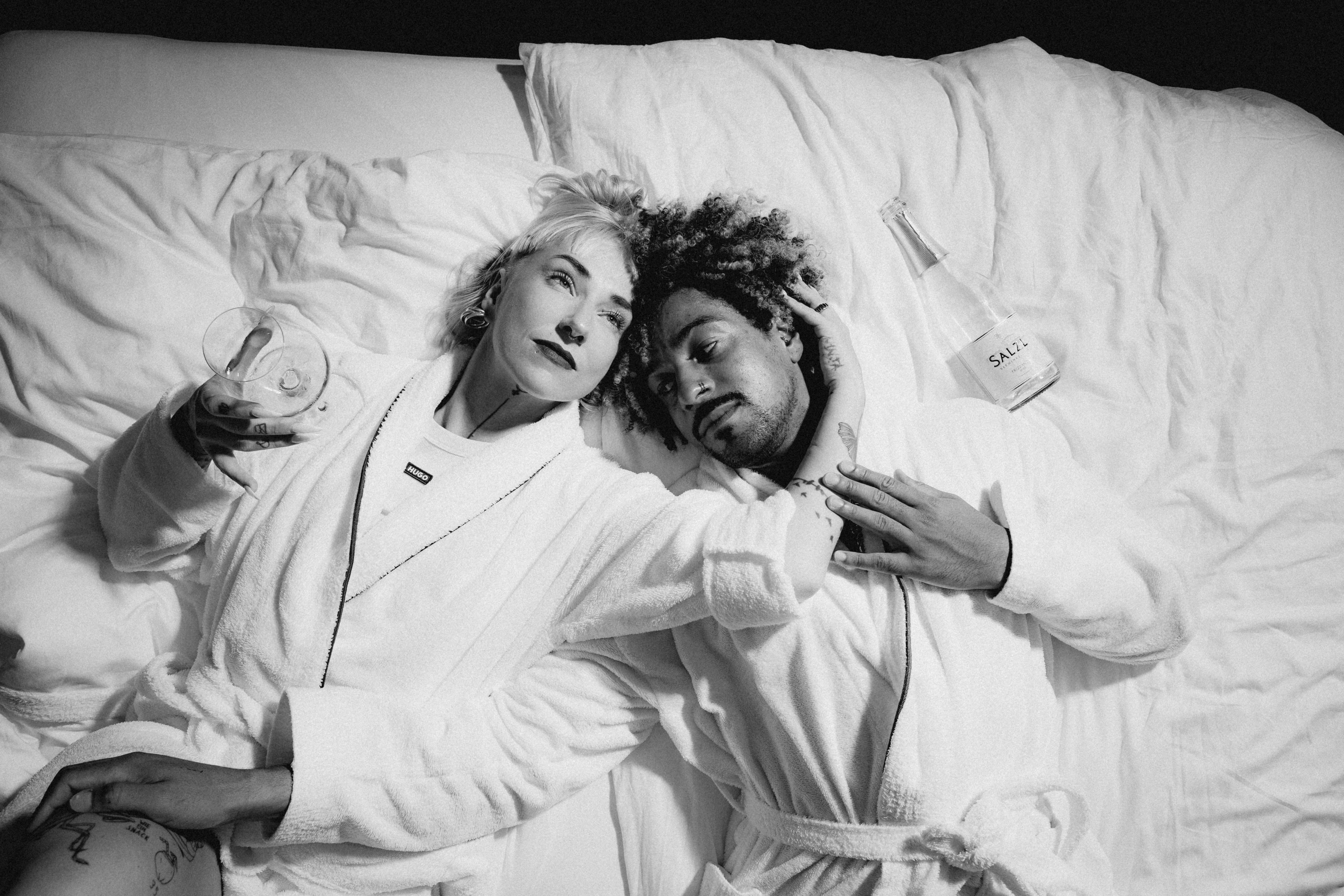 A black and white image of a neatly made bed, a bedside table, and a small trash can beside it.