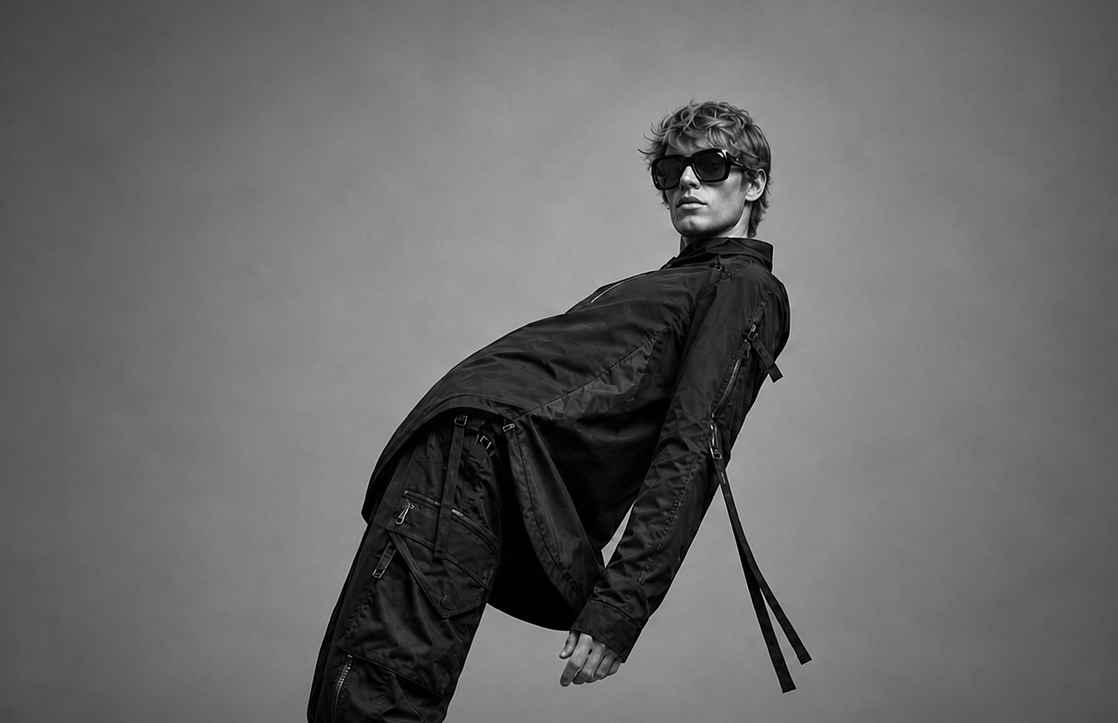 Black-and-white image of a person wearing sunglasses and dark clothing, leaning backward at an angle against a plain gray background.