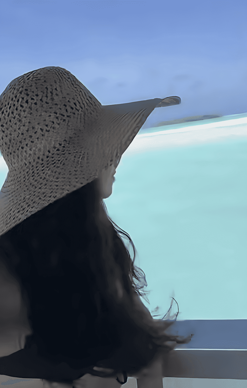 Woman with long dark hair and a sun hat looking out over blue ocean, a beach and blue sky
