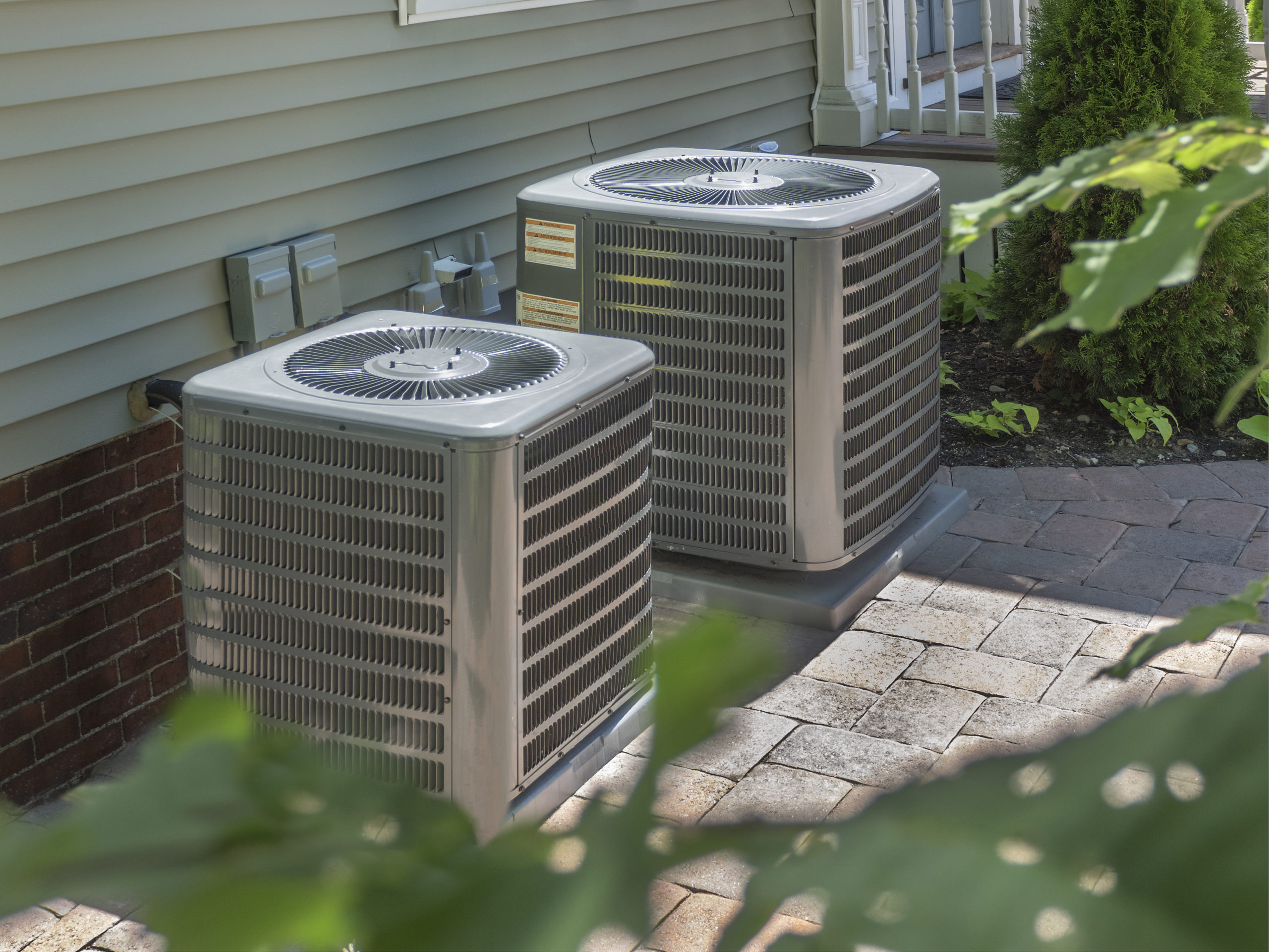 Two outdoor air conditioning units sit side by side next to a house with beige siding, showcasing a recent AC installation in Asheville. The units are on a paved surface near a brick foundation and surrounded by green foliage. A small staircase with white railings is partially visible in the background.