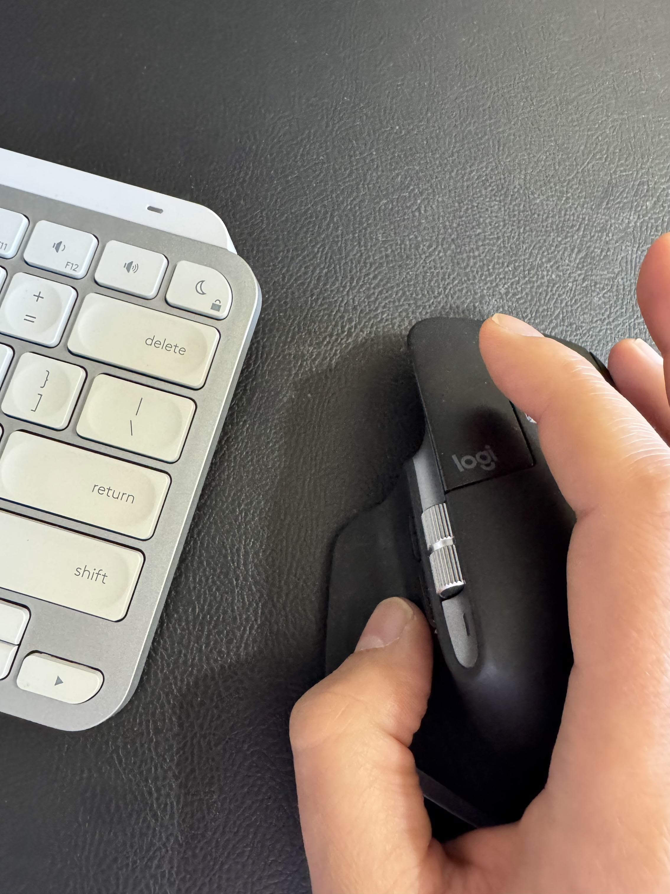 A close-up view of a hand using a black Logitech computer mouse on a dark surface, next to a silver wireless keyboard. The scene captures a minimalist and modern workspace setup, emphasizing productivity and technology.