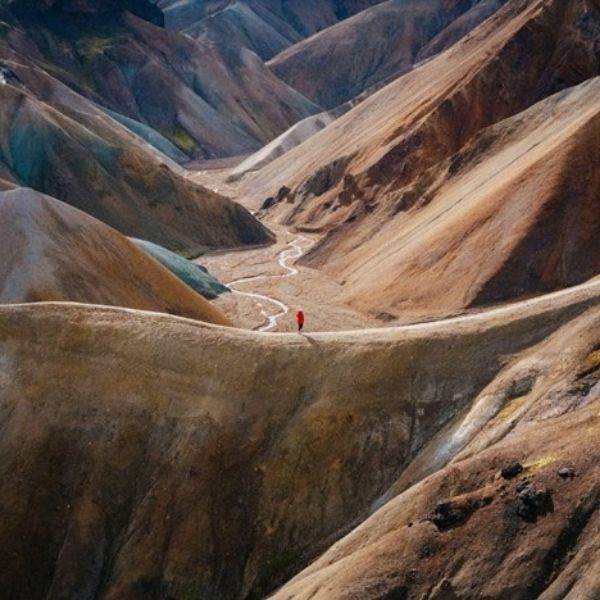  An aerial photo of the rainbow mountains of Landmannalaugar and the Torfajoköll volcanic system with a hiker in a red coat on the trail overlooking a river. The rhyolitic lava shaped the mountains that formed from the Torfajökull central volcanic eruptions.