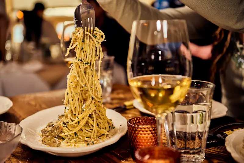 A person lifting a forkful of pasta over a table with a glass of white wine.