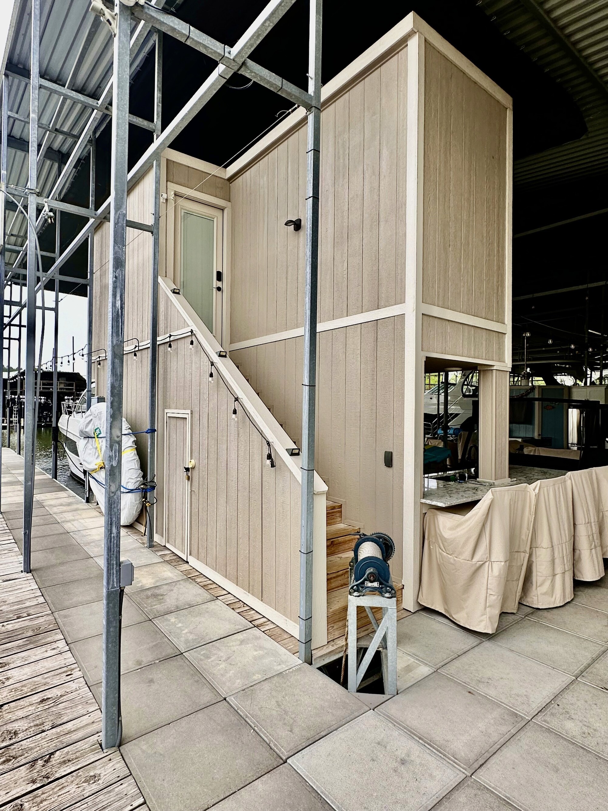 A beige wooden boathouse with a staircase and door is situated on a dock, surrounded by metal scaffolding and covered in protective canvas sheets.