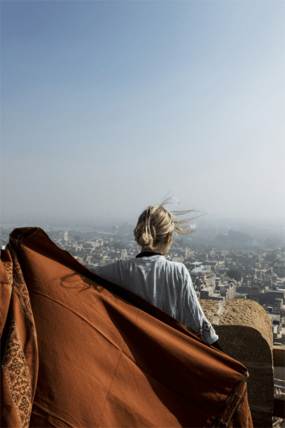 A person sits on a ledge, gazing at a hazy cityscape, with a warm-toned fabric draped beside them.