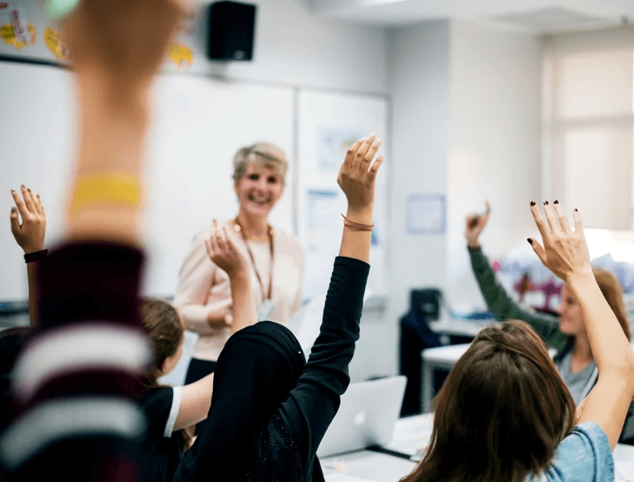 A classroom scene with a teacher standing at the front near a whiteboard, while several students seated at desks raise their hands. Laptops and classroom materials are visible, with daylight coming in through windows.