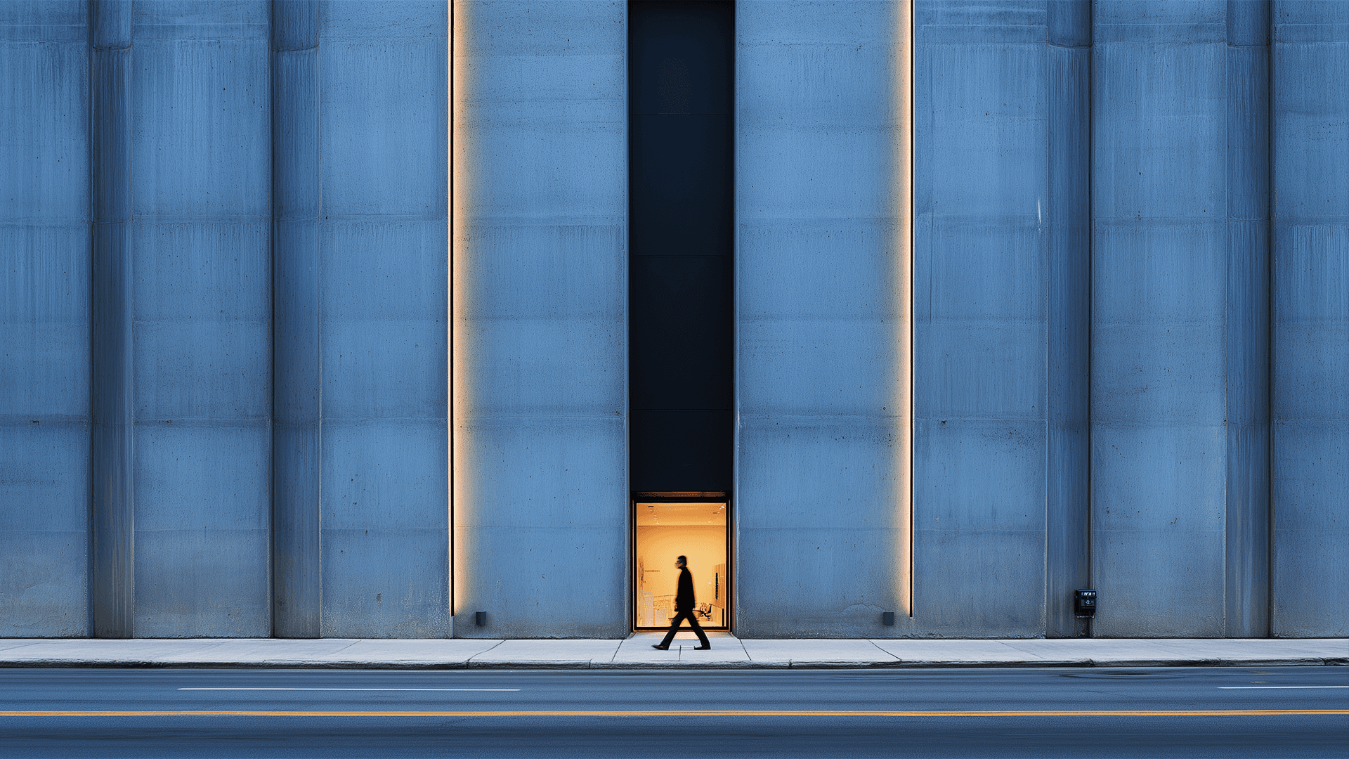 Fotografia simétrica de uma fachada arquitetônica imponente em concreto azul-acinzentado. No centro, uma faixa vertical escura desce até uma porta iluminada com uma luz amarela quente. A silhueta de uma pessoa caminha apressada pela calçada em frente à entrada, criando um contraste entre a escala humana e a monumentalidade do edifício