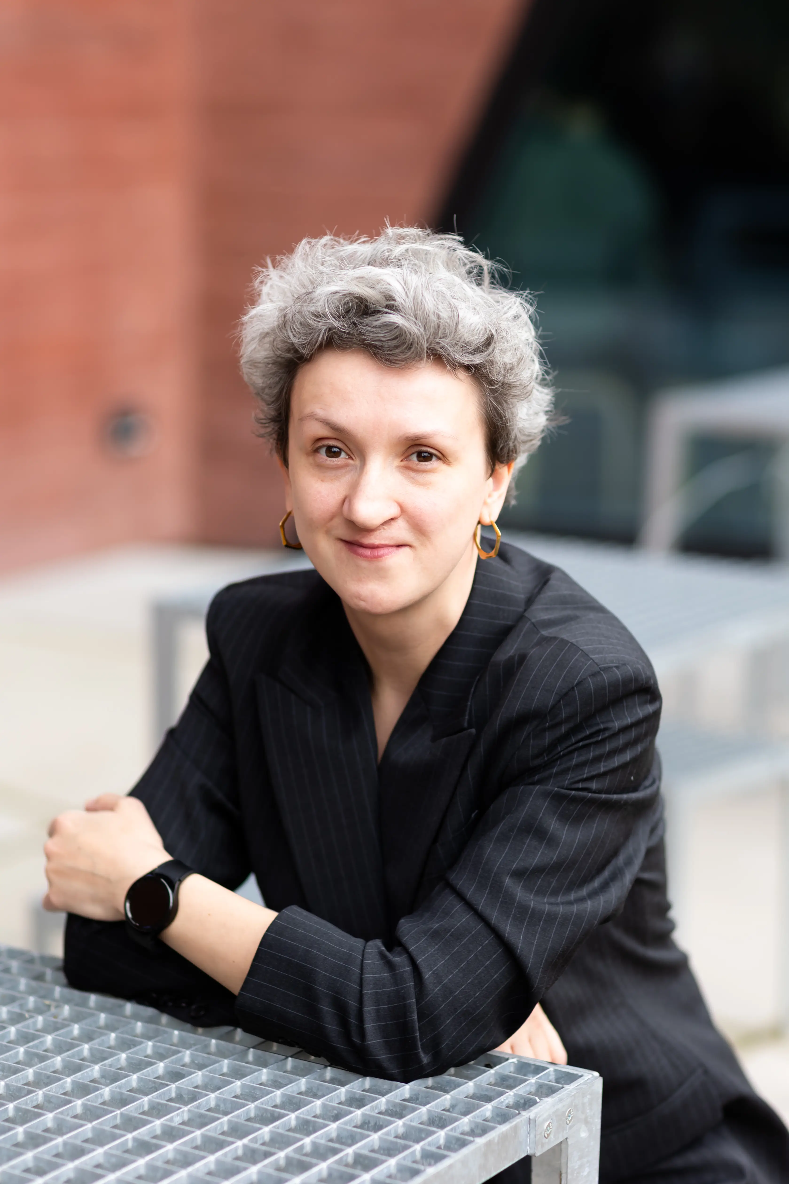 Kobieta o krótkich, kręconych, siwych włosach, ubrana w ciemną marynarkę w prążki i złote kolczyki w kształcie kółek, siedzi przy metalowym stole na świeżym powietrzu i patrzy w stronę kamery z lekkim uśmiechem.