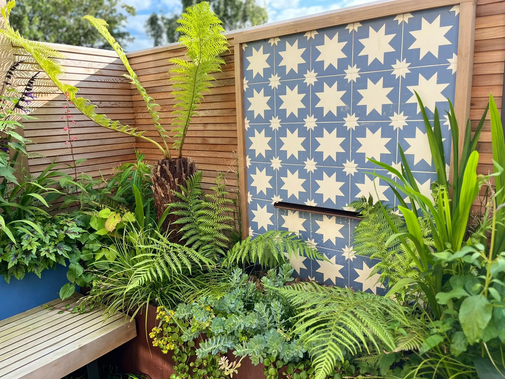 A lush garden with green plants and a textured wall featuring a blue and white patterned design.