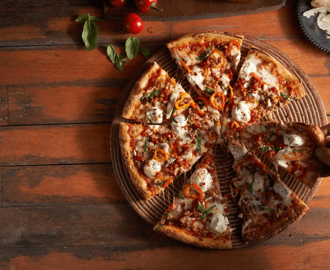 A top-down view of a supreme pizza on a warm handmade pizza stone on a wood table and a white man’s hand picking up one of the pizza slices. Other ingredients are included in the shot.