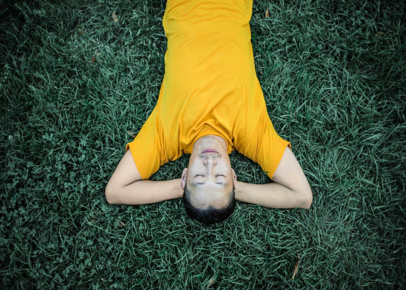 Upside down image of smiling man in yellow shirt laying down on grass with his arms crossed behing his head