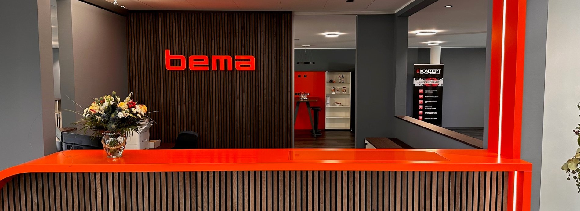 Bema Firmenlogo an Empfangstheke. Moderner Empfangsbereich mit Blumen und roter Beleuchtung. Holzdekor, Büroeingang