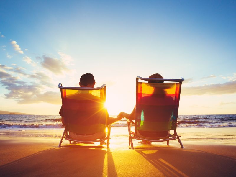 Two people are seated in colorful beach chairs on the sand, holding hands and watching a stunning sunset over the ocean. The sun casts a warm glow across the horizon, with rays of light peeking through the silhouetted figures, creating a tranquil and romantic scene.