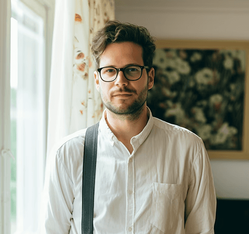 A man with glasses and a beard stands indoors, wearing a white shirt and suspenders. He looks calm by a window with floral curtains and a floral painting.