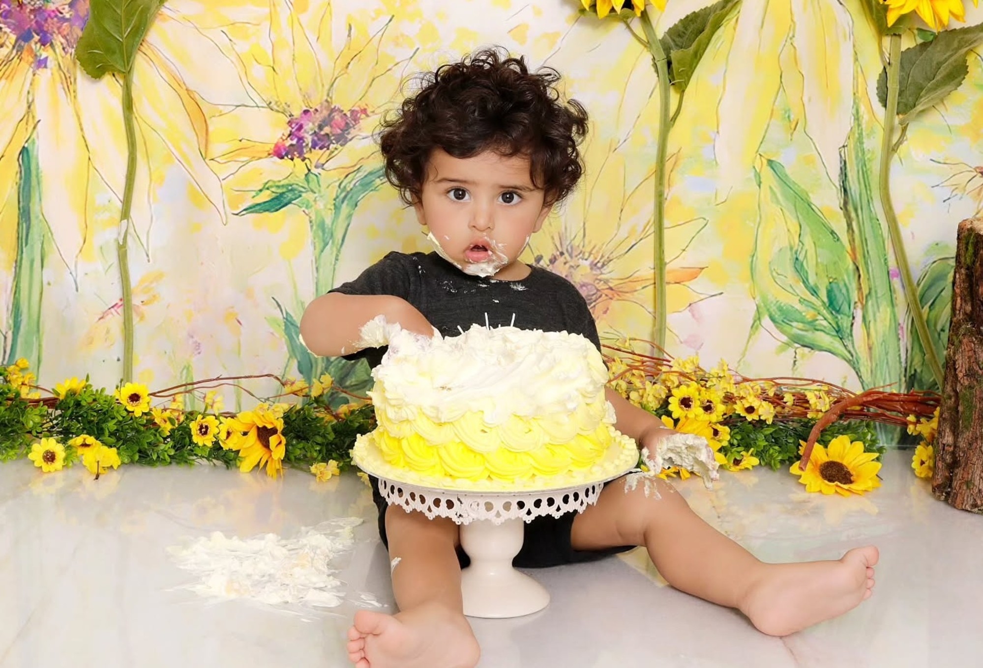 Baby with hands covered in smash cake frosting with a sunflower-themed background, during a Dubai session at Mahvin Photography.
