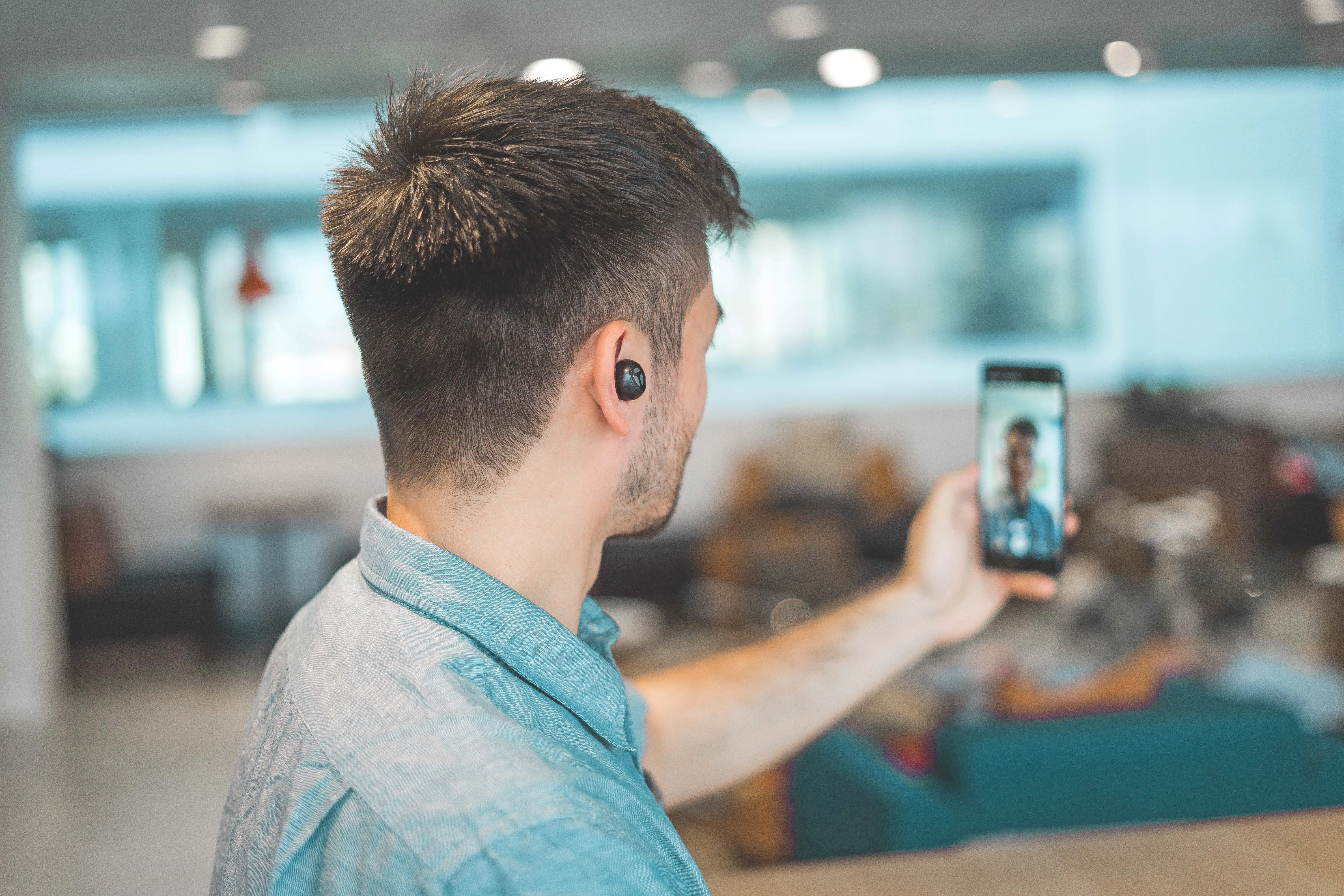 Man wearing wireless earbuds taking a selfie or video call with a smartphone in a modern office setting