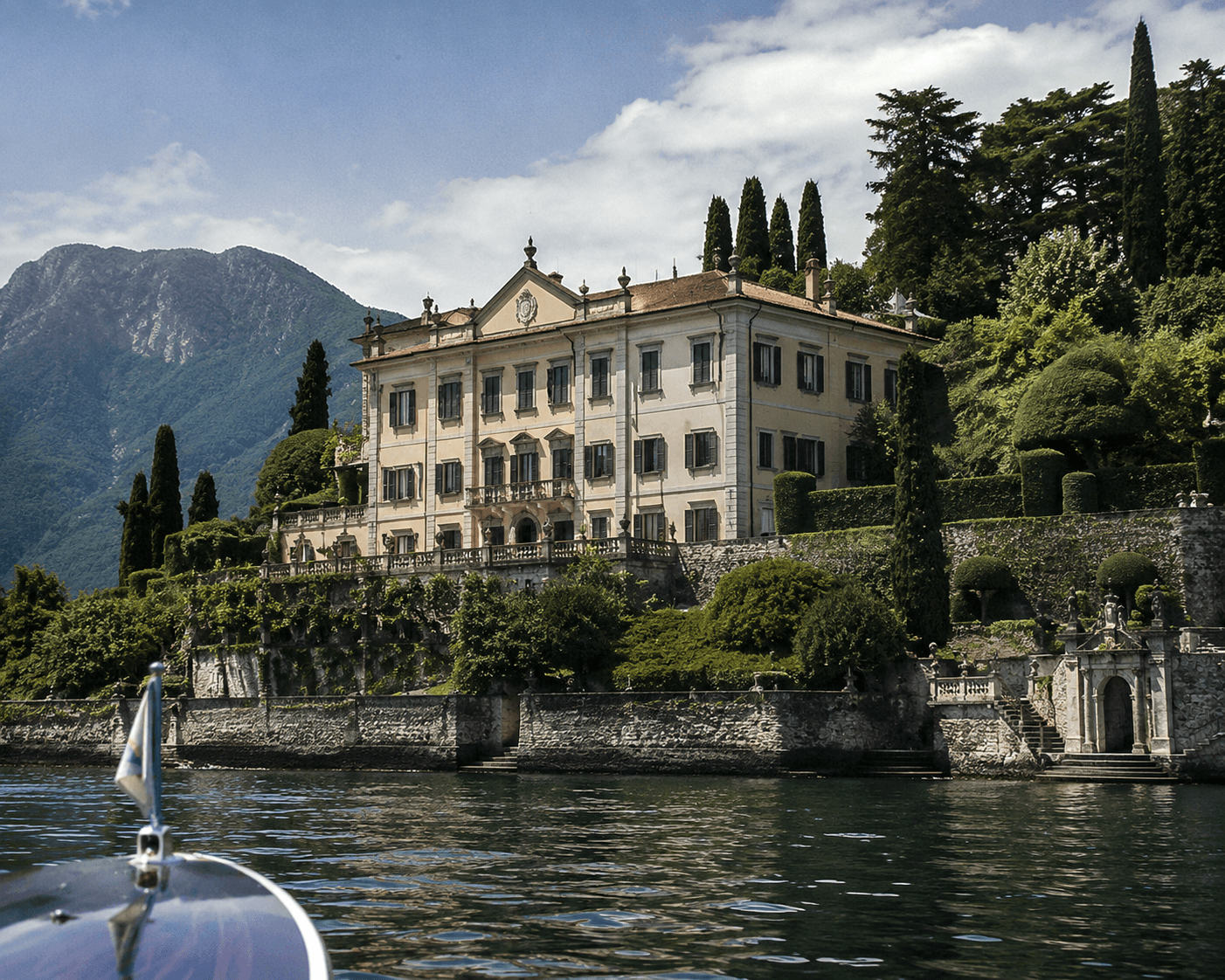 A low-angle photograph taken from the bow of a wooden boat looking up at a grand, historic Italian villa situated on the edge of Lake Como. The multi-story beige estate features dark window shutters, ornamental roof finials, and stone balustrades set above terraced gardens filled with manicured green hedges, cypress trees, and stone archways. Dramatic blue mountains and a bright blue sky with light clouds form the backdrop across the shimmering water.